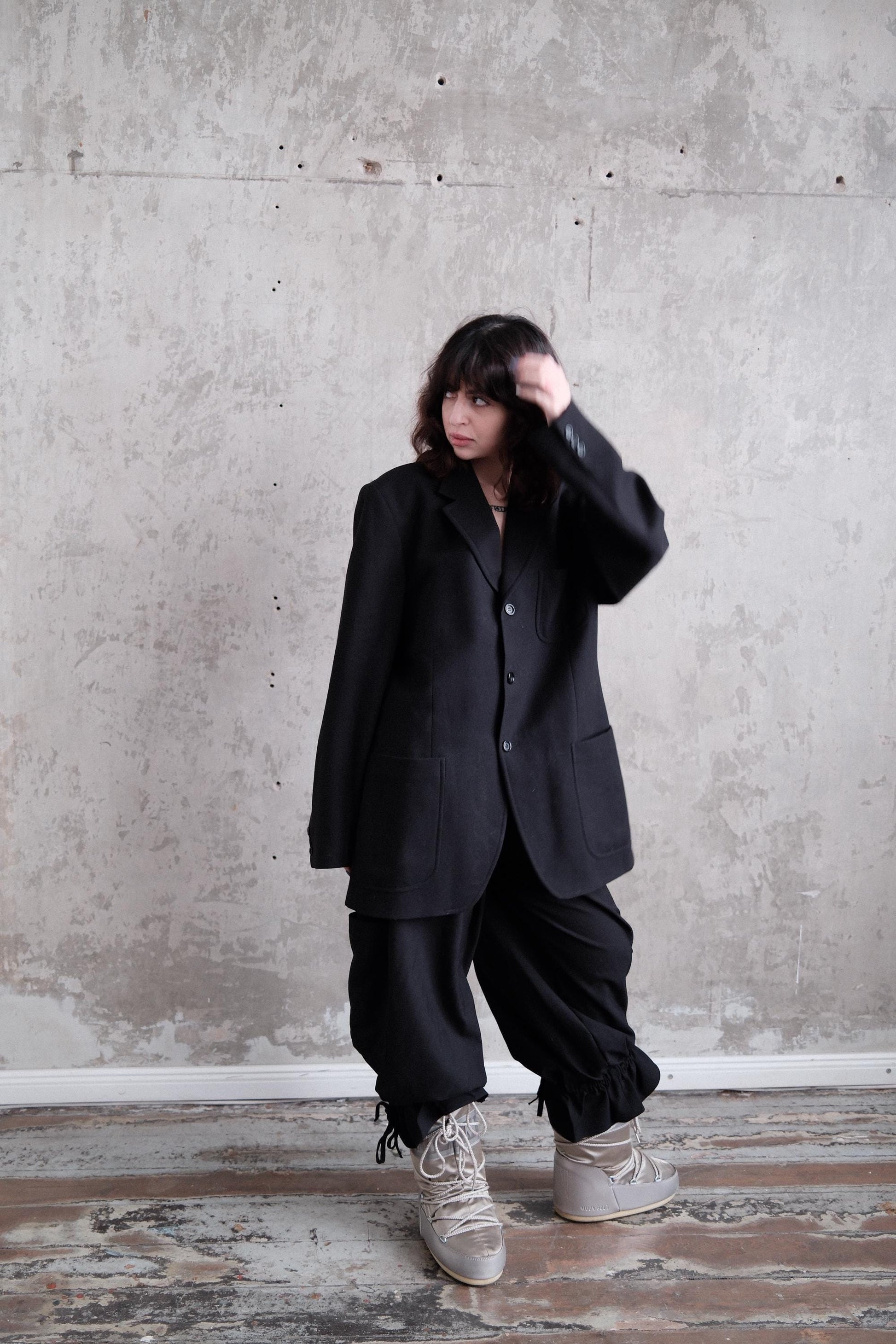 A person wearing a black jacket and pants stands in a room with a concrete wall.