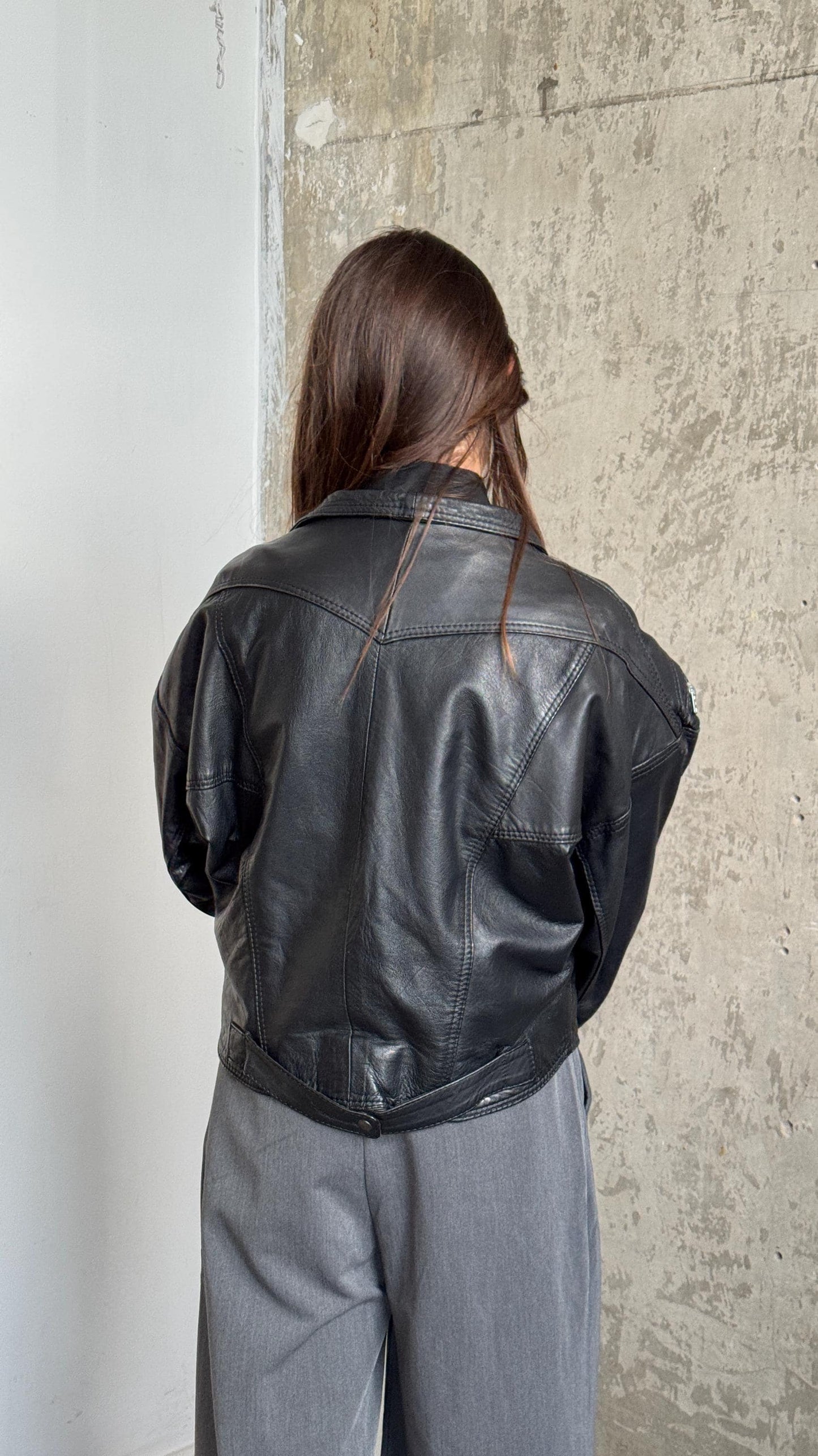A person wearing a black leather jacket stands in front of a concrete wall.