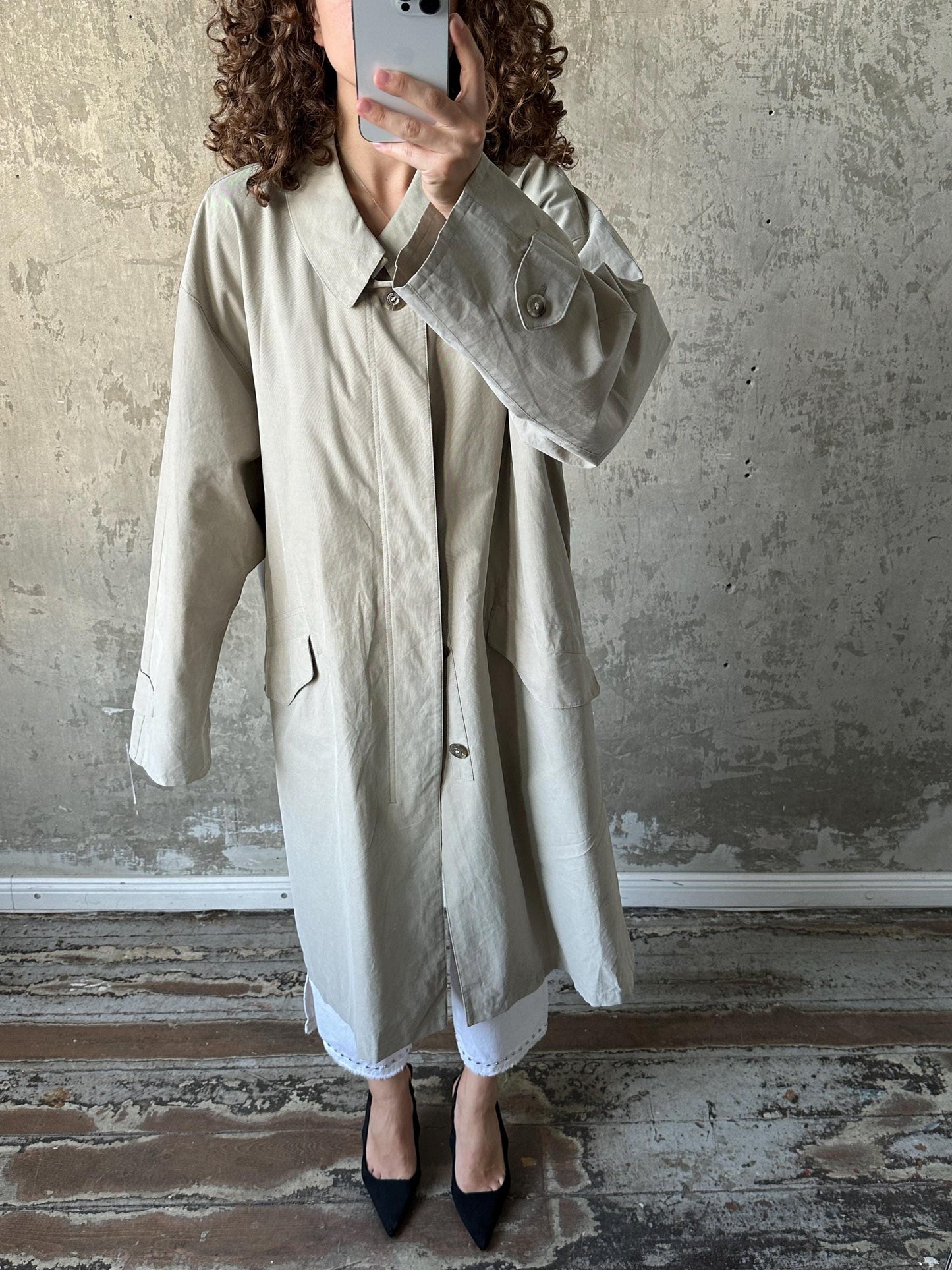 A person is taking a selfie with a smartphone, wearing a beige coat and black shoes, standing in front of a concrete wall.