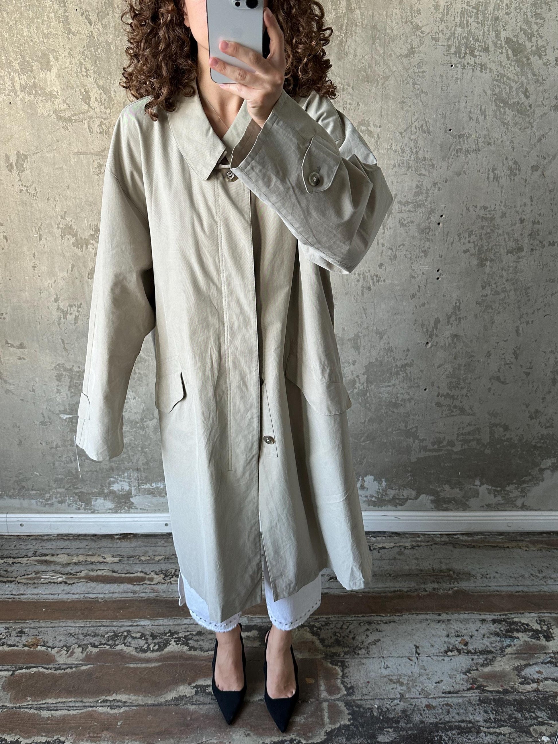 A person is taking a selfie with a smartphone, wearing a beige coat and black shoes, standing in front of a concrete wall.