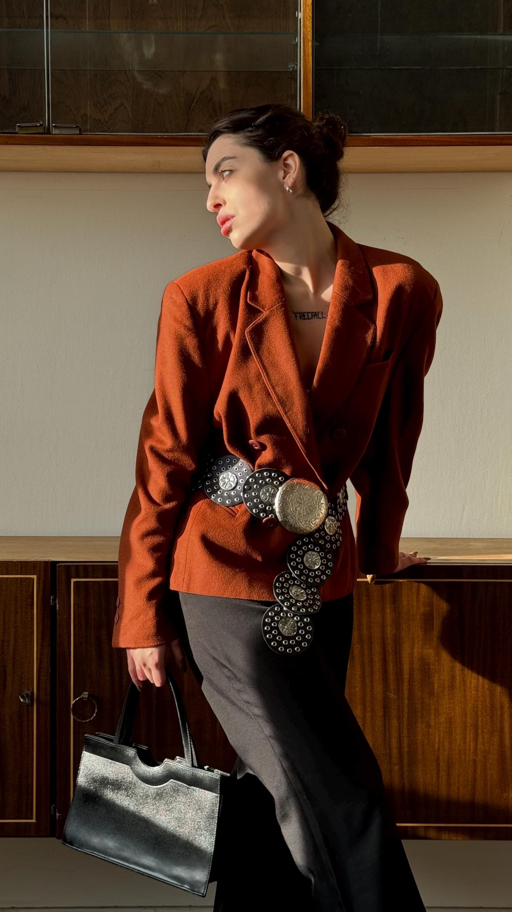 A woman wearing a red blazer and carrying a black purse stands in front of a wooden cabinet.