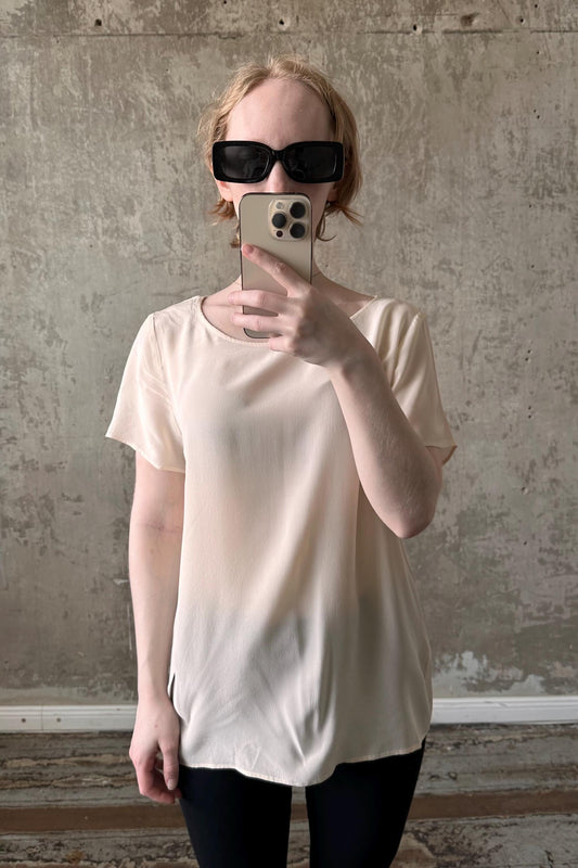 A person is taking a selfie with a smartphone, wearing a white top and sunglasses, against a concrete wall background.