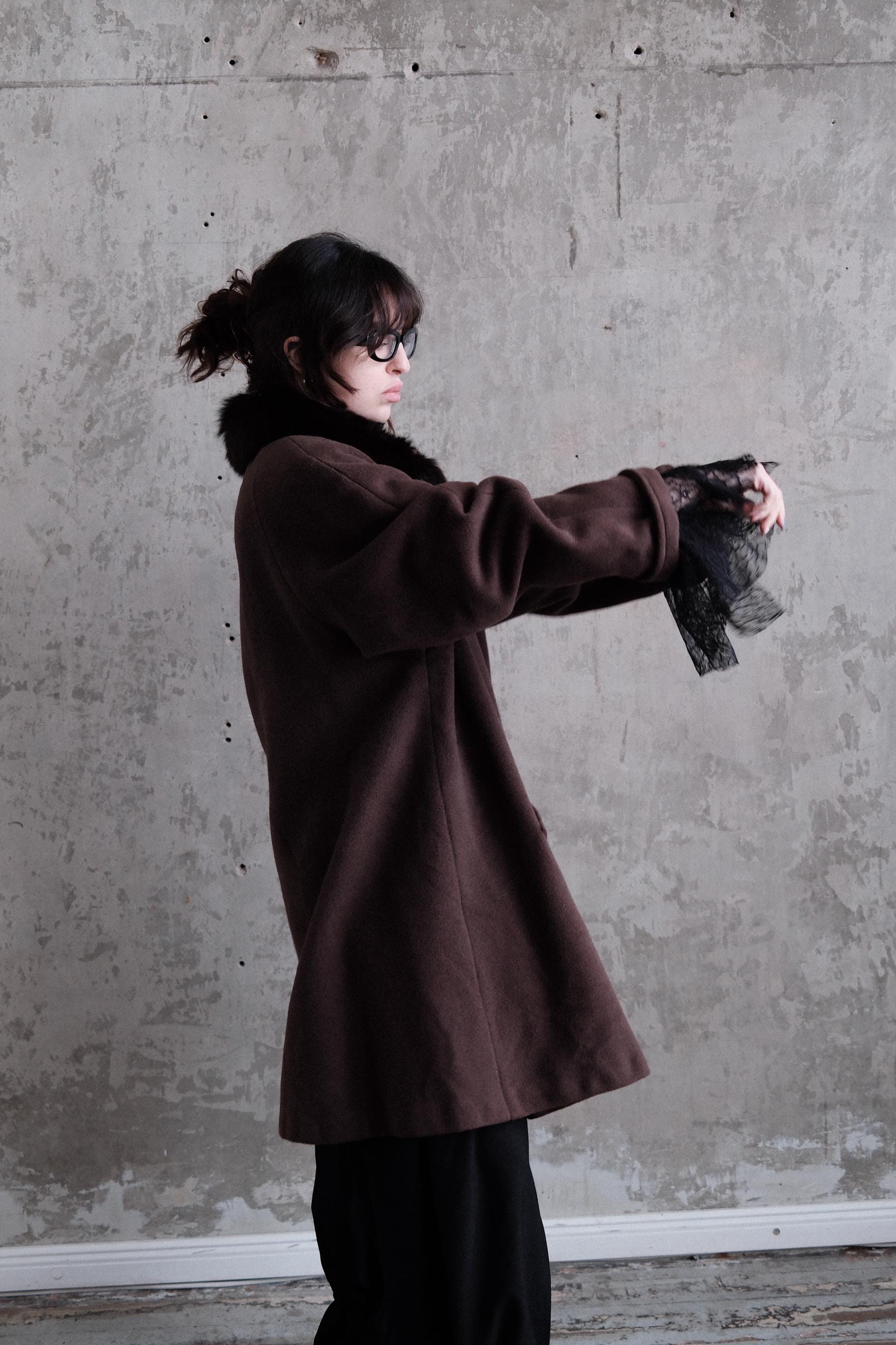 A person wearing a brown coat and black pants is standing against a concrete wall.