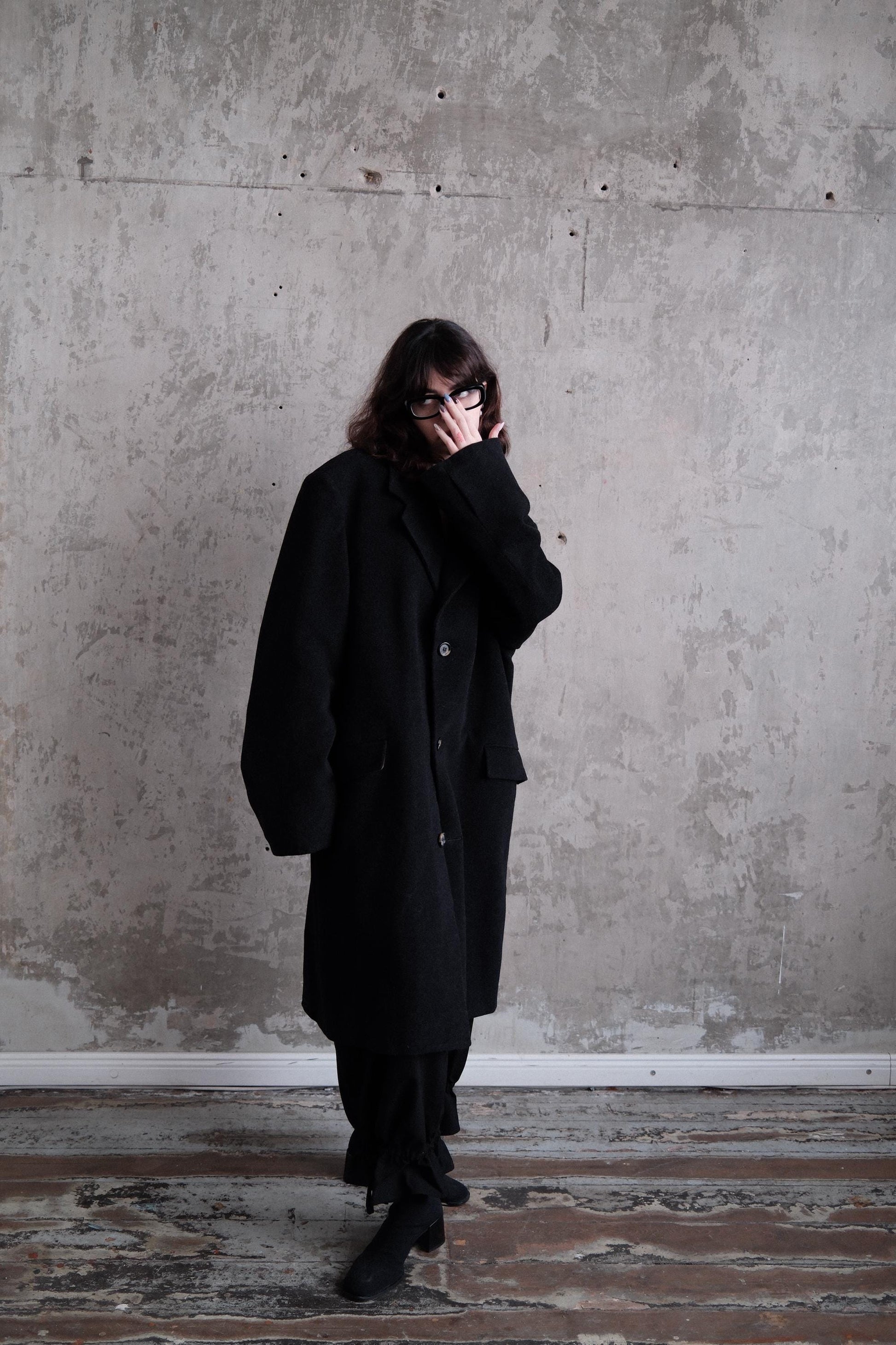 A person wearing a black coat stands in a room with a concrete wall.