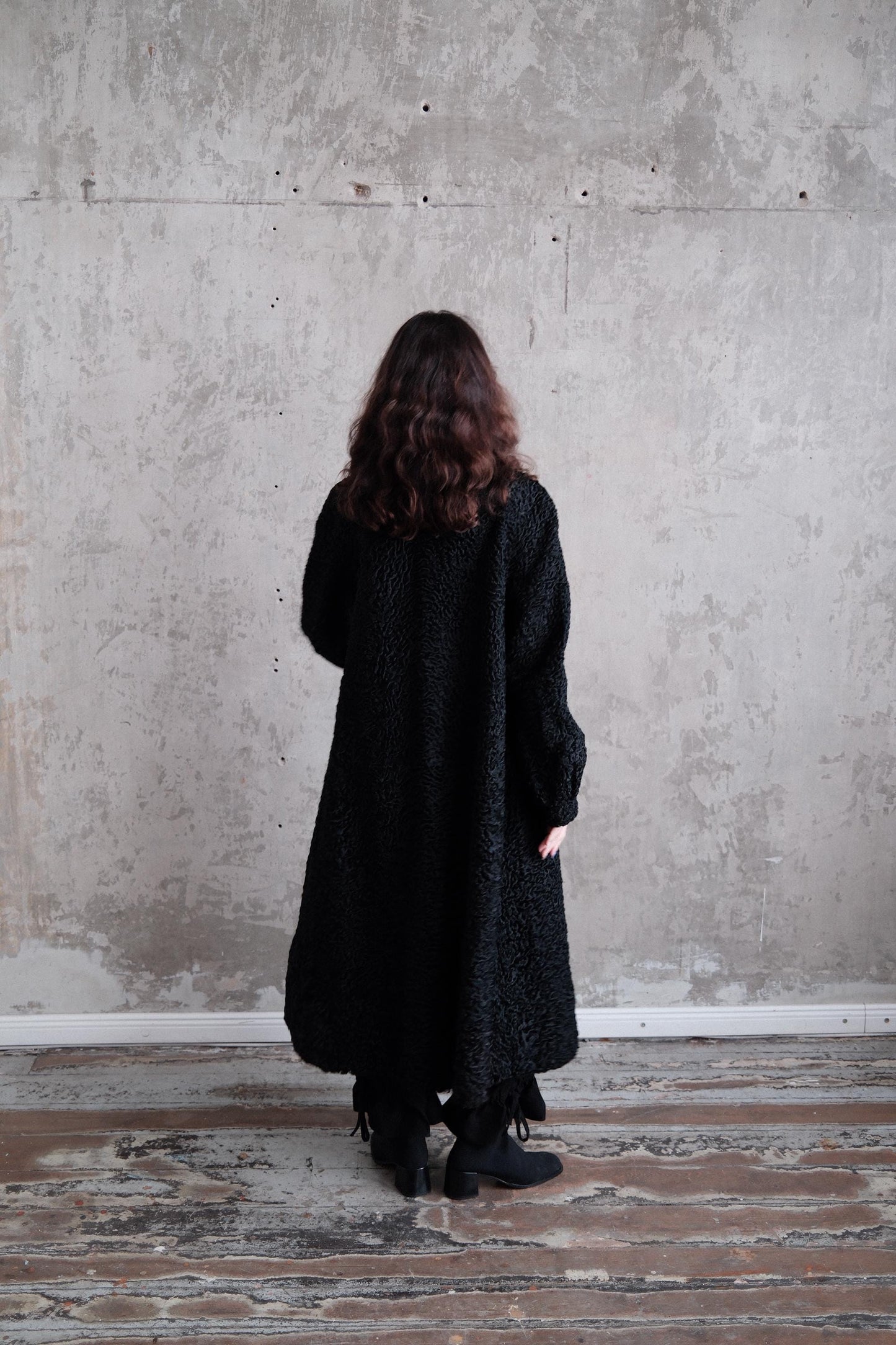 A person wearing a black coat stands in a room with a concrete wall and wooden floor.