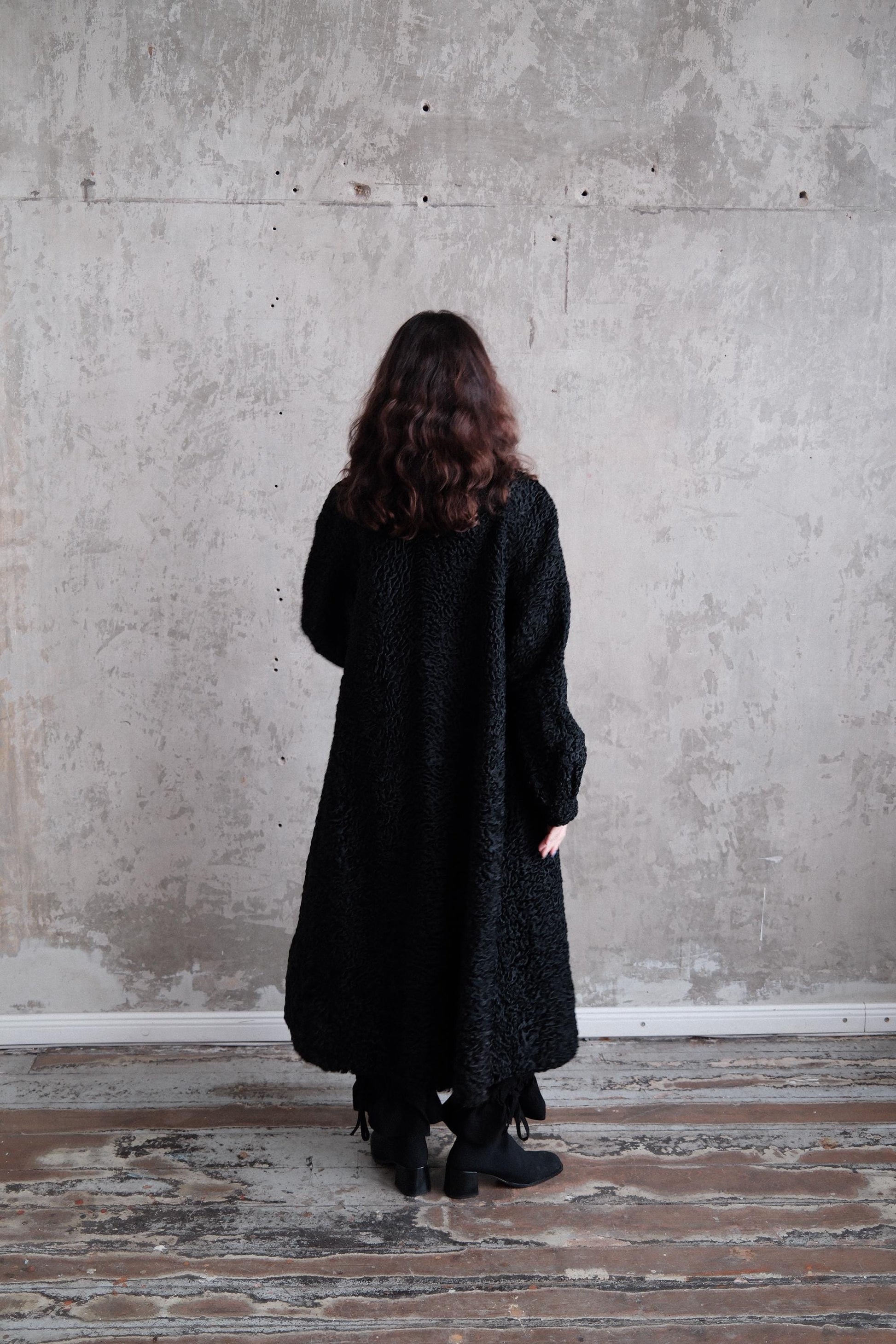 A person wearing a black coat stands in a room with a concrete wall and wooden floor.
