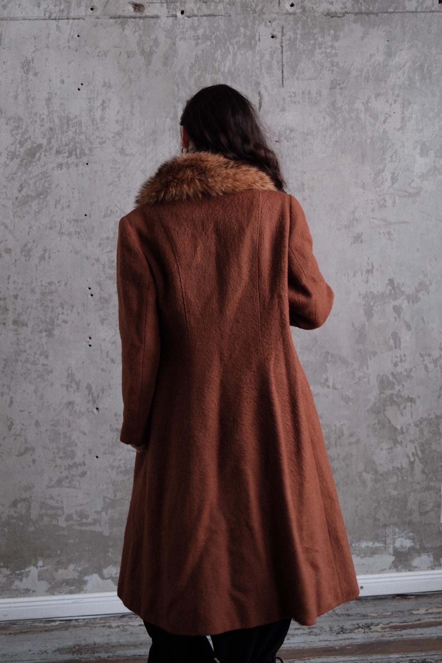 a person wearing a brown coat with a fur collar, standing against a concrete wall.