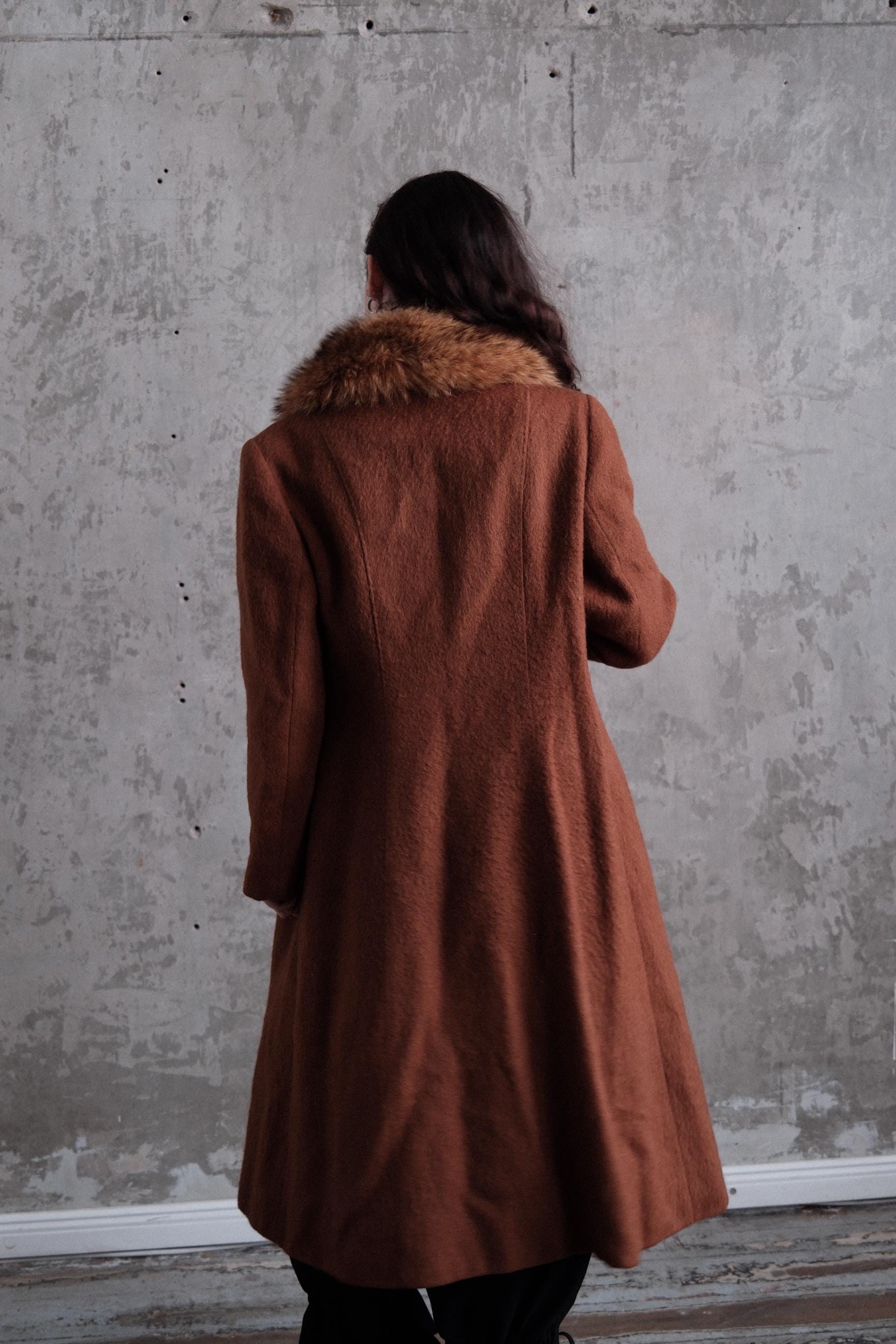 a person wearing a brown coat with a fur collar, standing against a concrete wall.