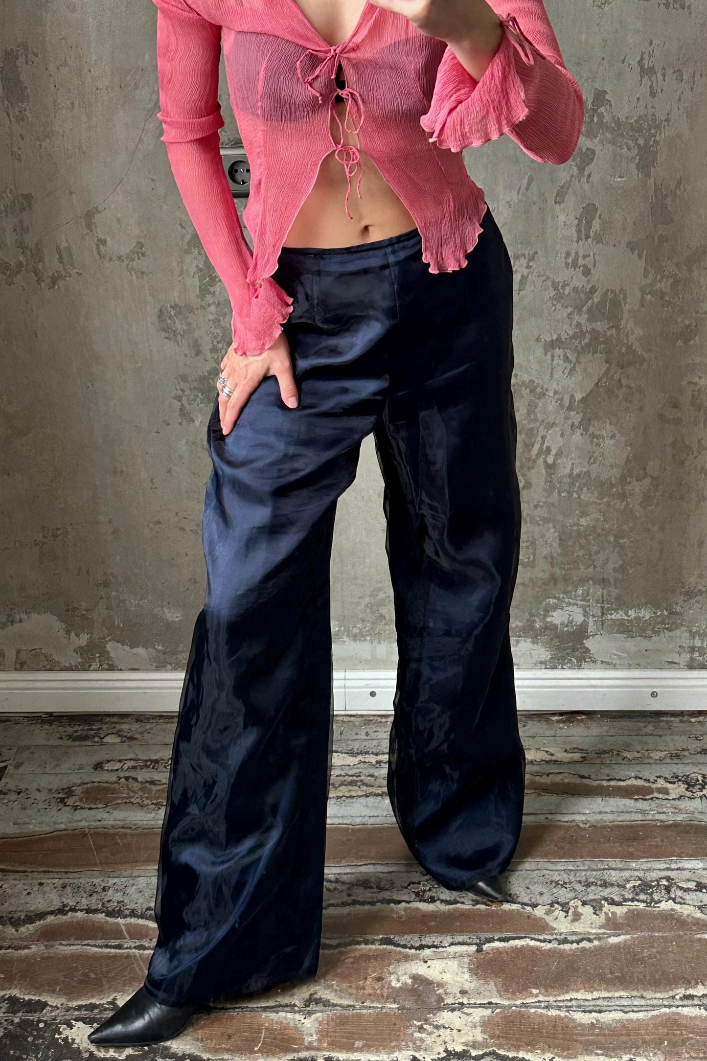 A person wearing a pink top and navy blue pants is standing in front of a concrete wall.