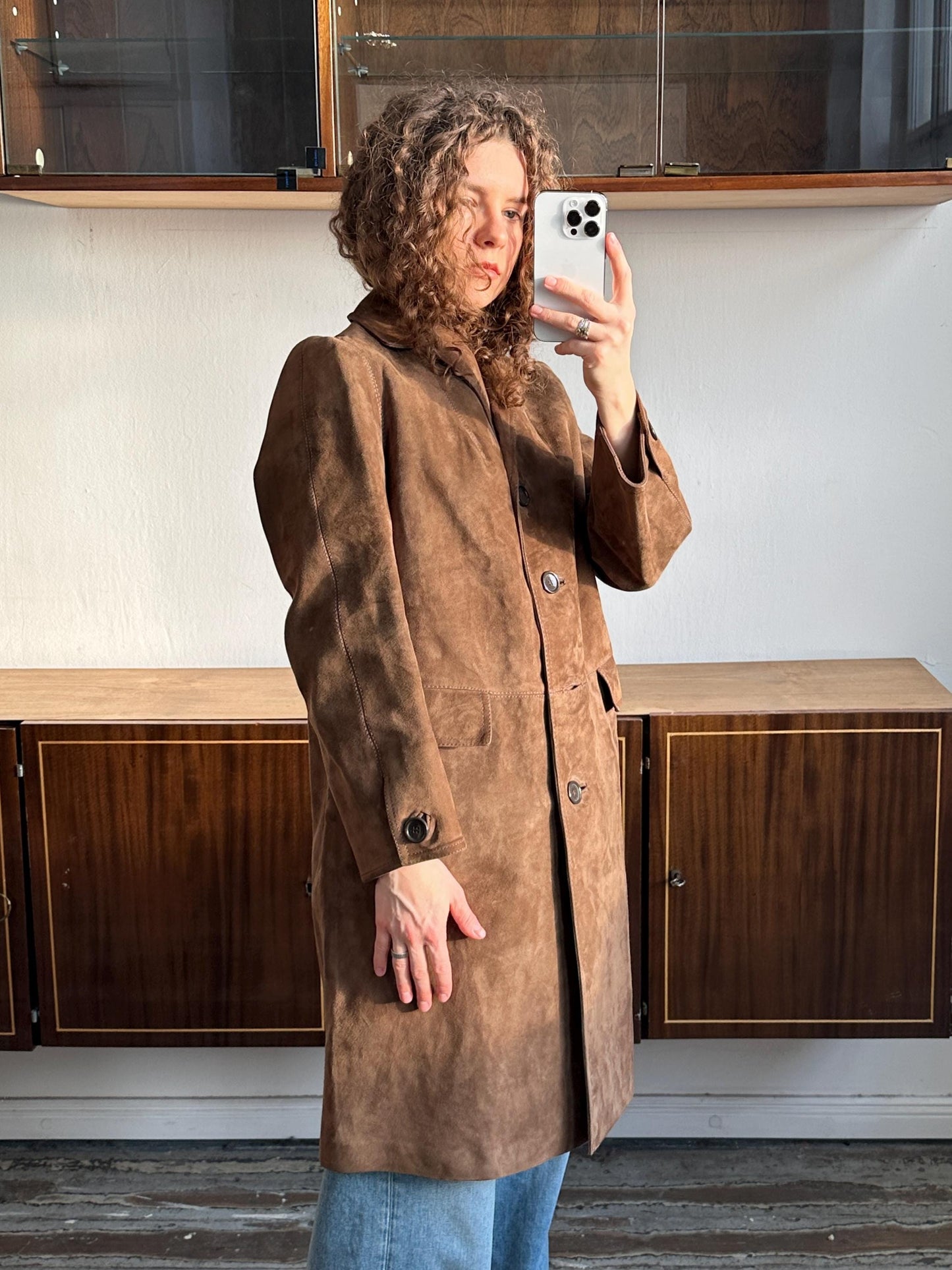 A woman with curly hair is taking a selfie with a white iPhone in front of a wooden cabinet.