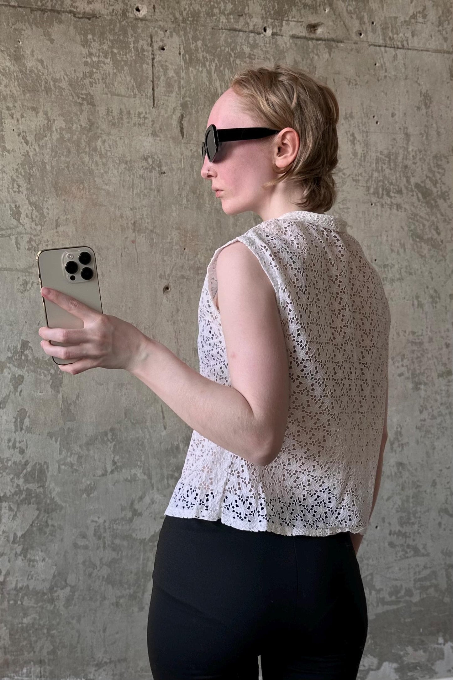 A woman wearing a white sleeveless top and sunglasses is holding a smartphone and taking a selfie in front of a concrete wall.