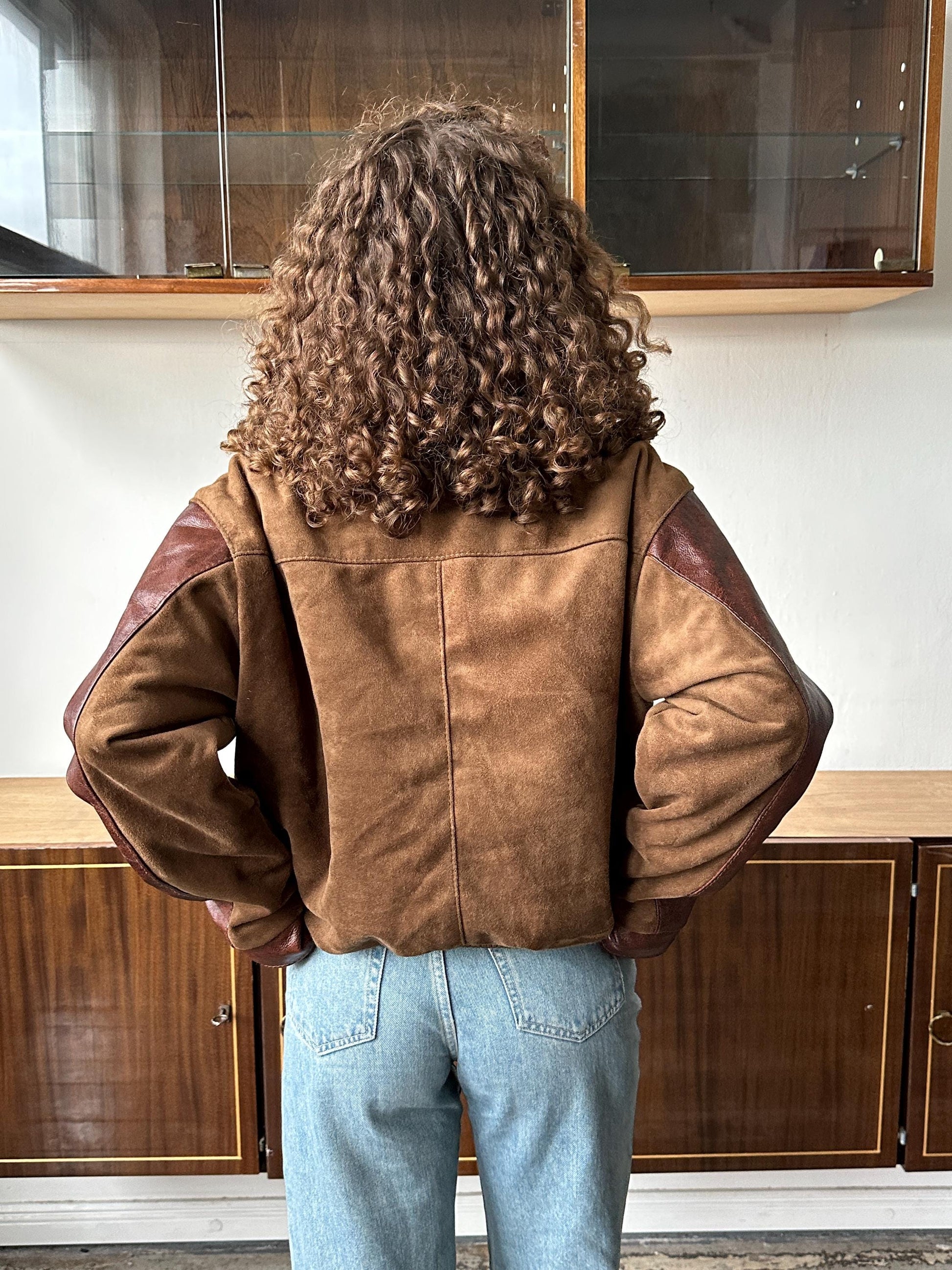 a person with curly hair wearing a brown jacket and blue jeans, standing in front of a wooden cabinet.