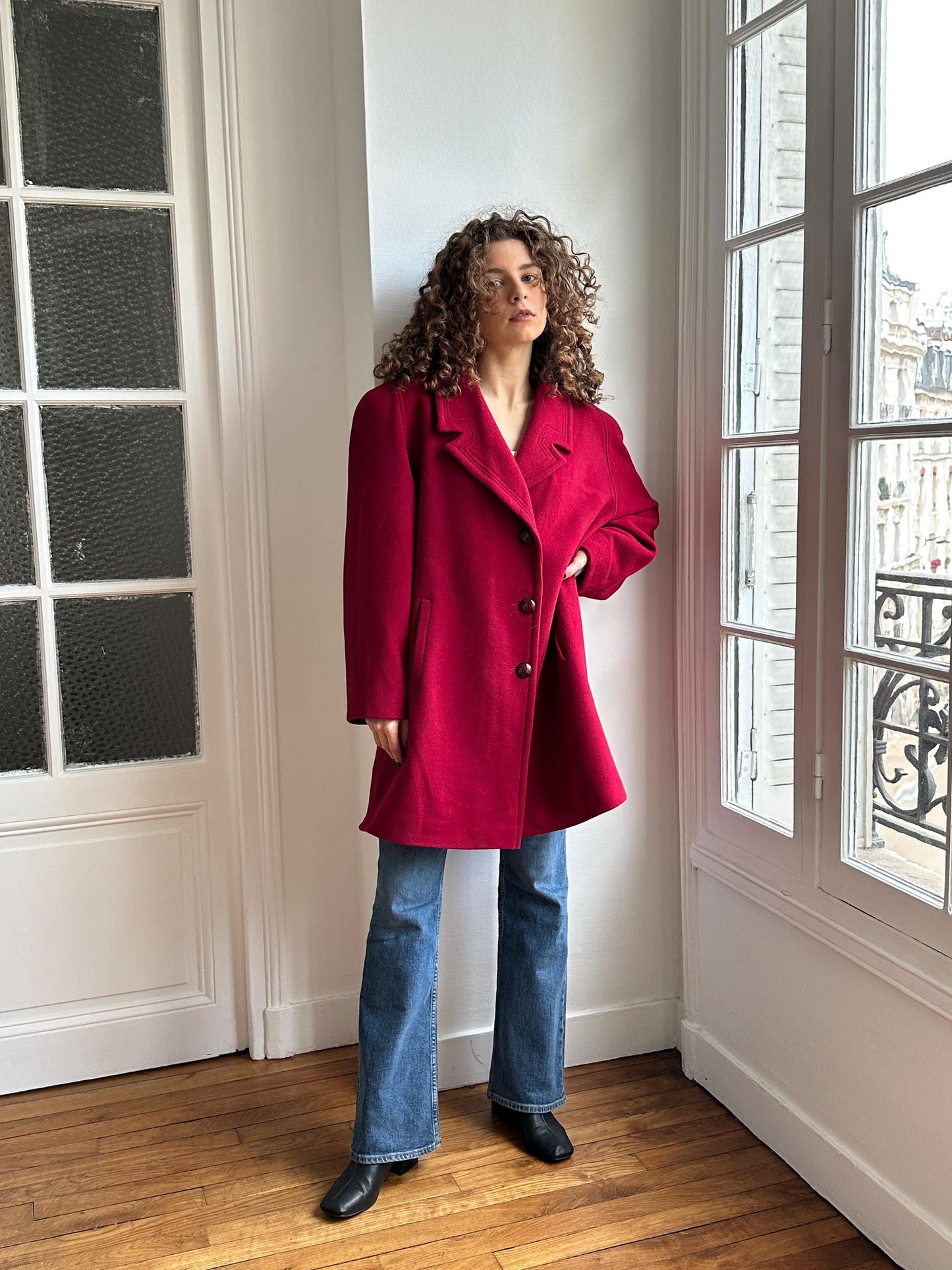 A person wearing a red coat and blue jeans is standing in a room with a window and a white wall.