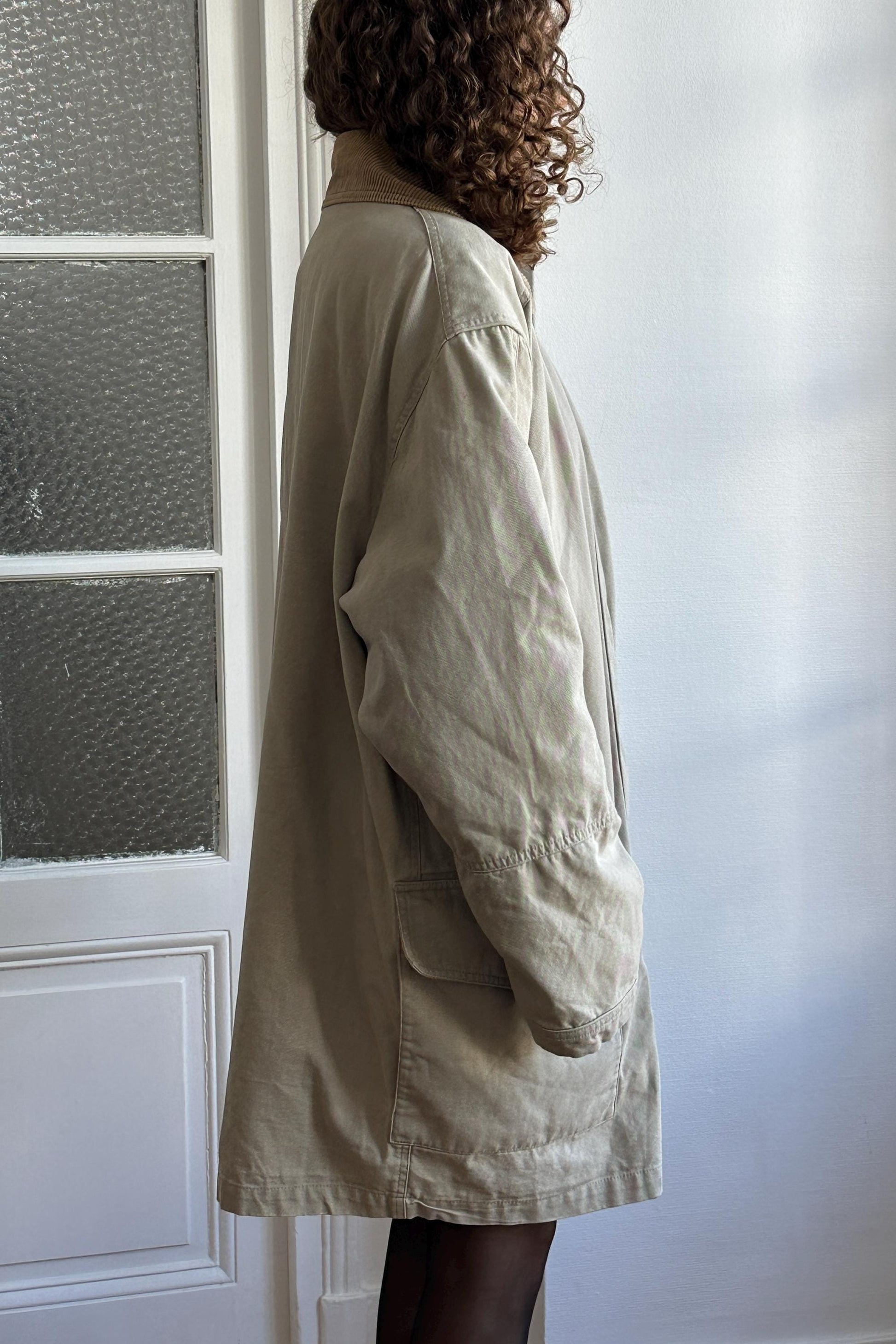 A person wearing a beige coat stands in front of a white door.