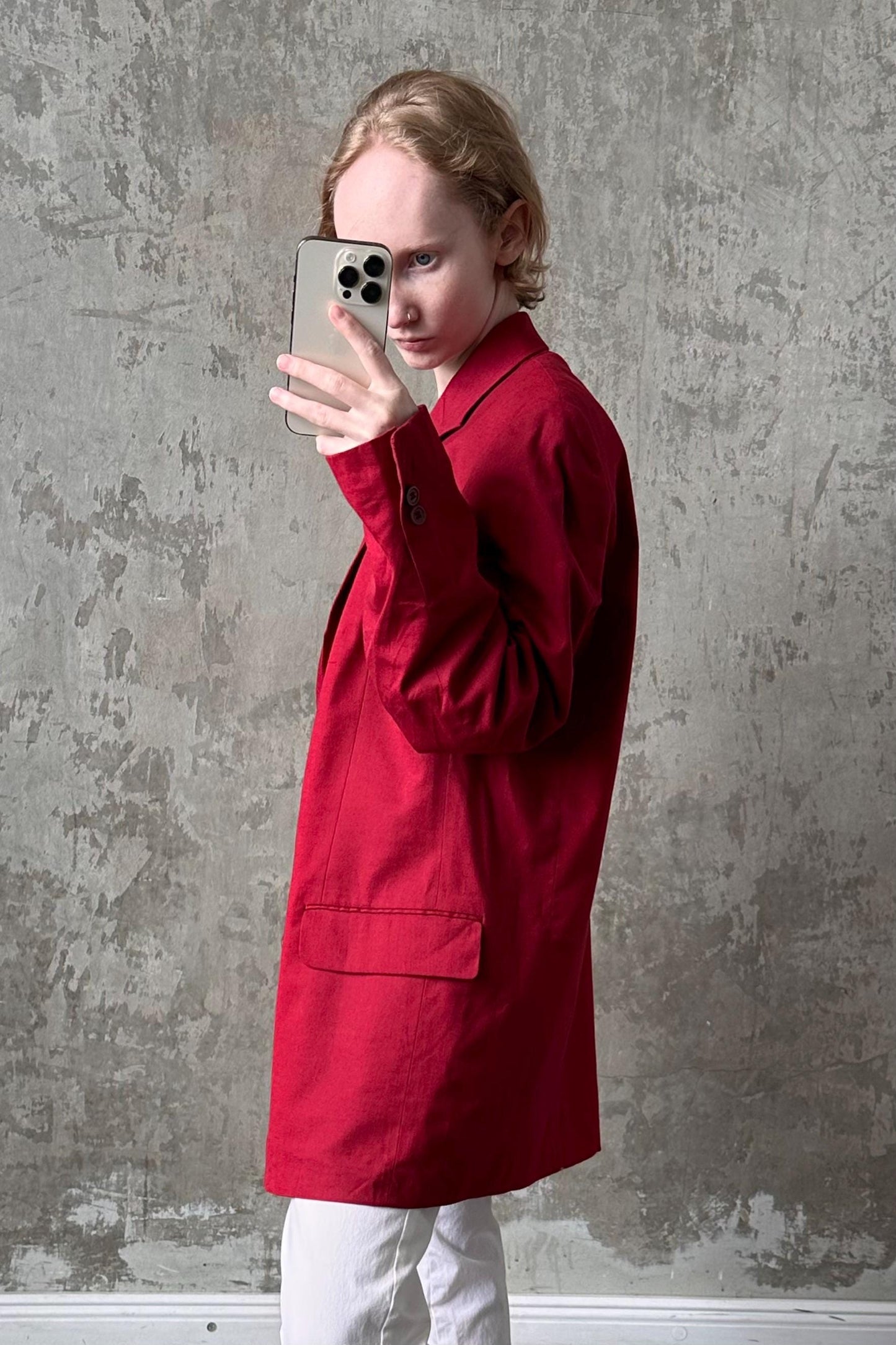 A person in a red coat is holding a smartphone up to their face, taking a selfie against a concrete wall.