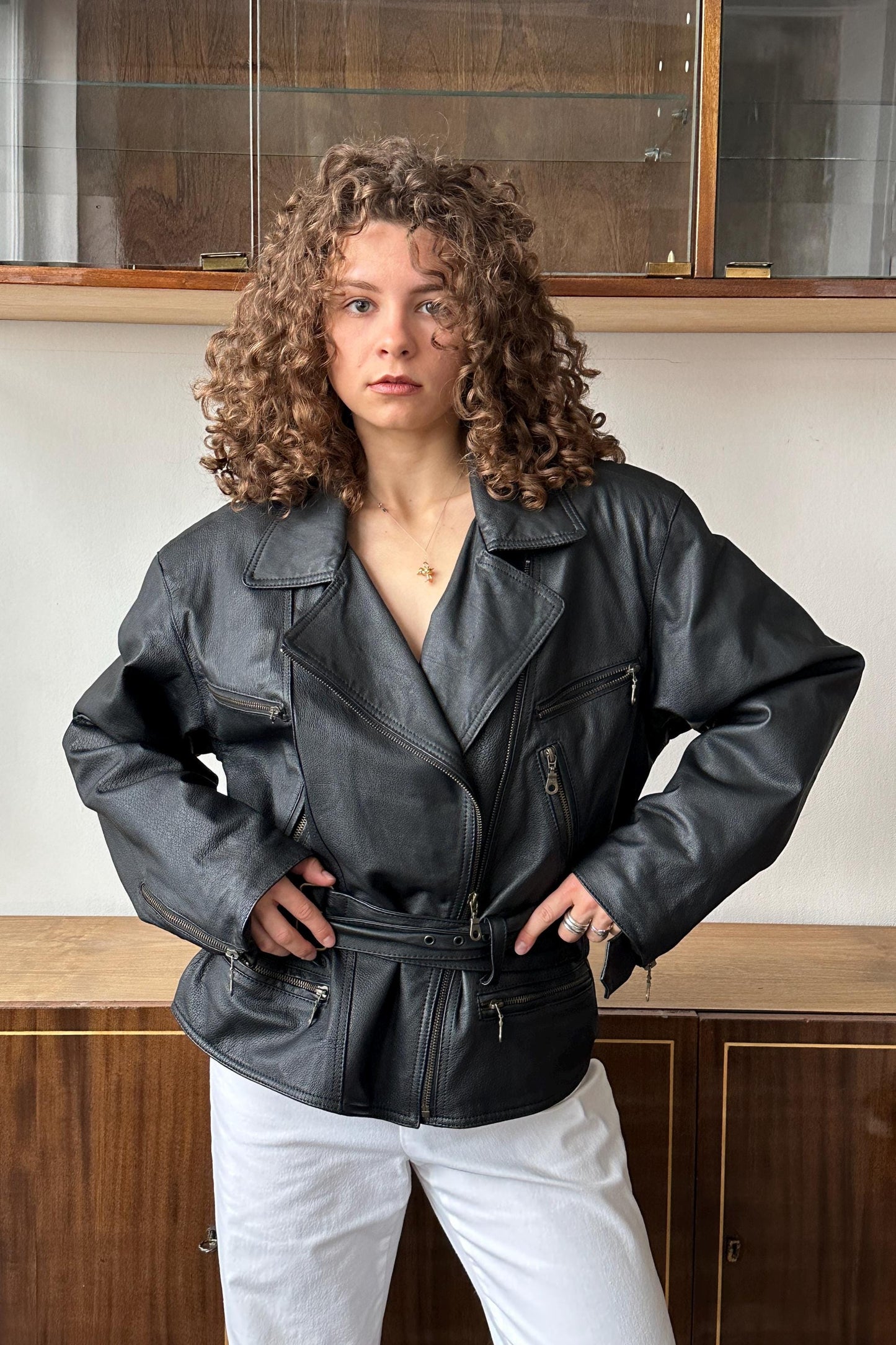 A woman with curly hair wearing a black leather jacket stands confidently in front of a wooden cabinet.