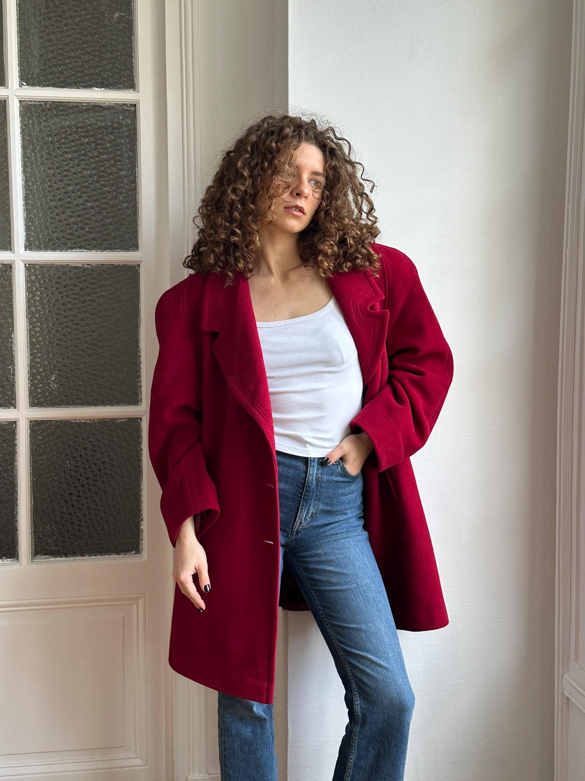 A woman with curly hair wearing a red coat and white top stands in front of a white door with glass panels.