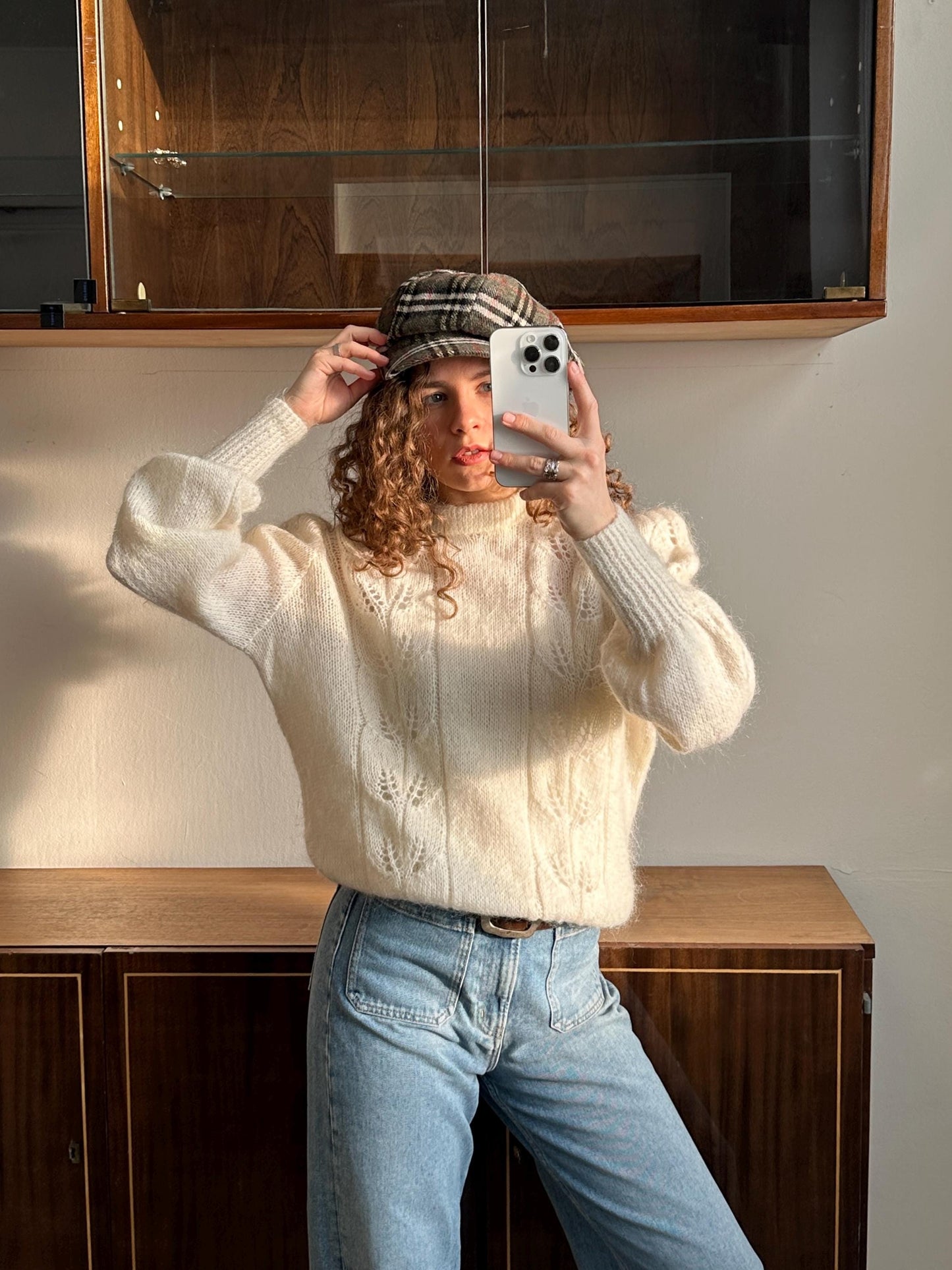 A woman with curly hair is taking a selfie with a white phone, wearing a white sweater and blue jeans.