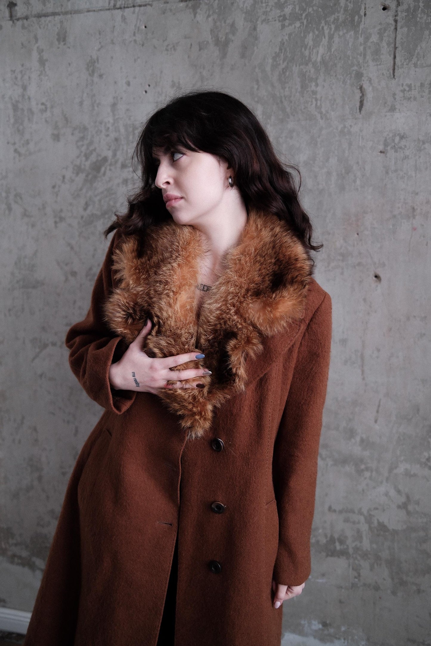 A woman wearing a brown fur coat is lying down on a concrete floor, looking up with a thoughtful expression.