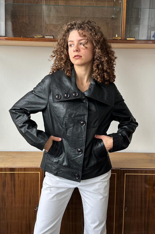 A woman with curly hair wearing a black leather jacket stands in front of a wooden cabinet.