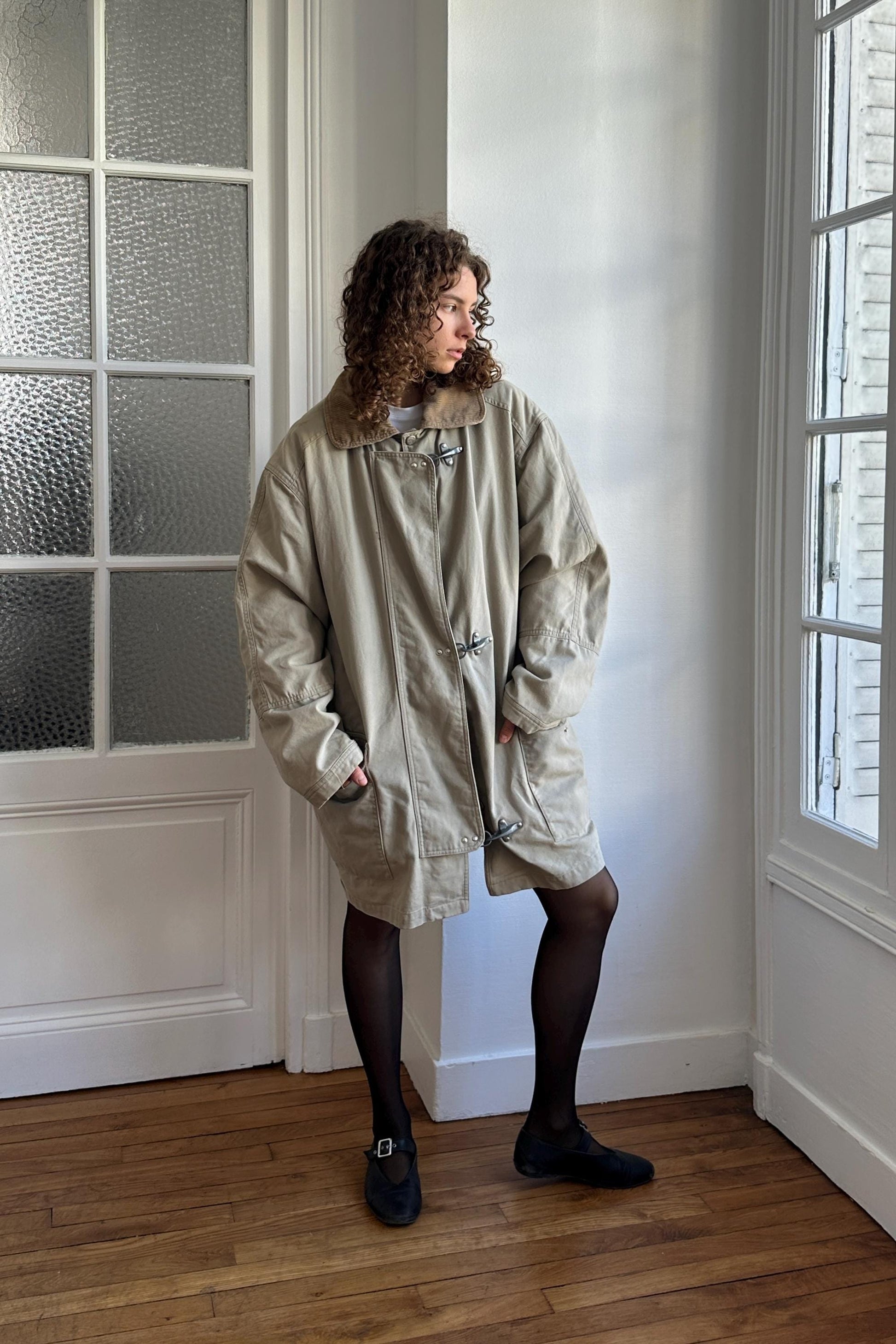 A person wearing a beige coat stands in a room with a window and a white door.
