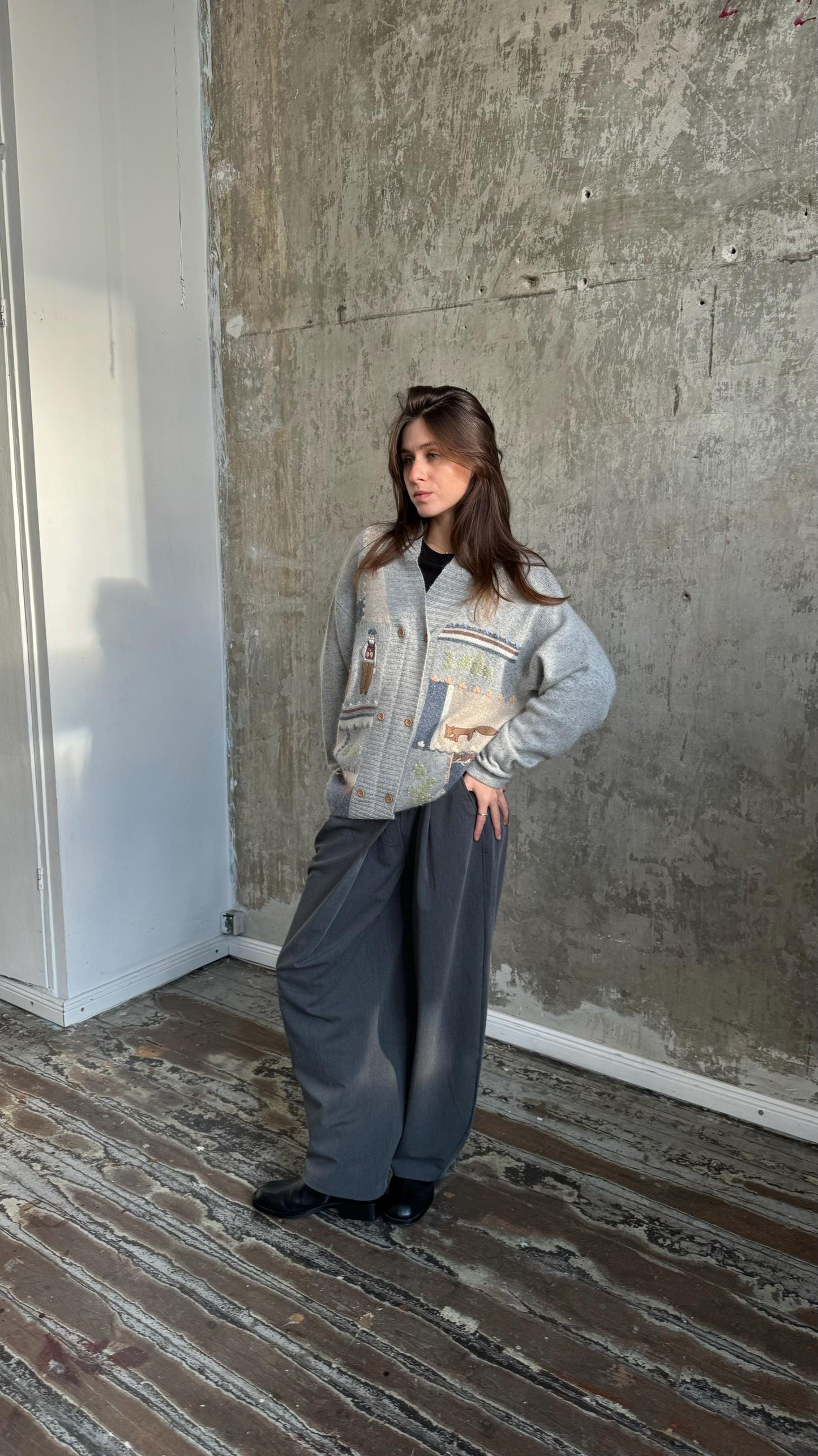 A woman wearing a gray sweater and gray pants stands in a room with a concrete wall.