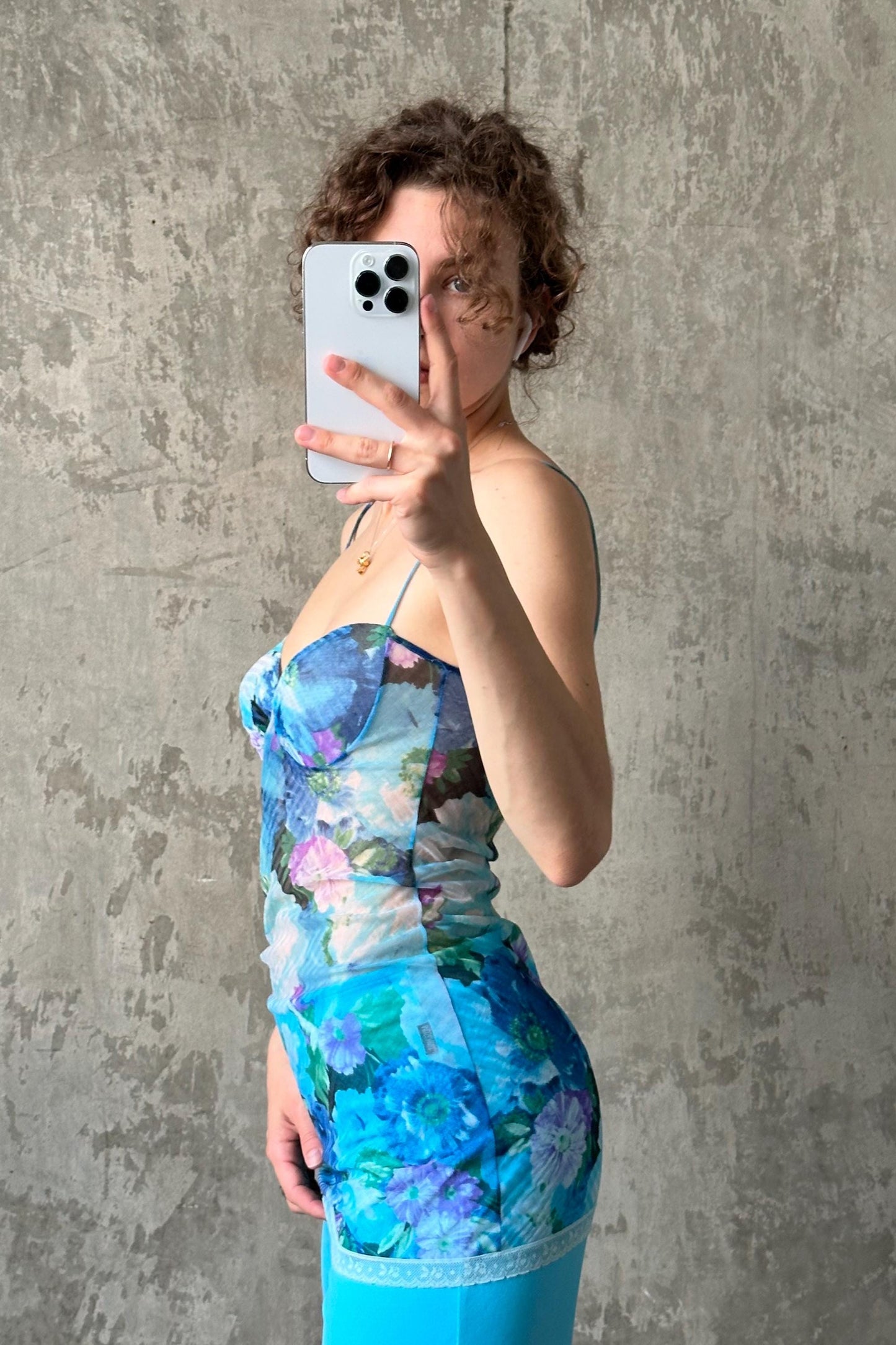 A woman in a blue floral dress is taking a selfie with a smartphone against a concrete wall.