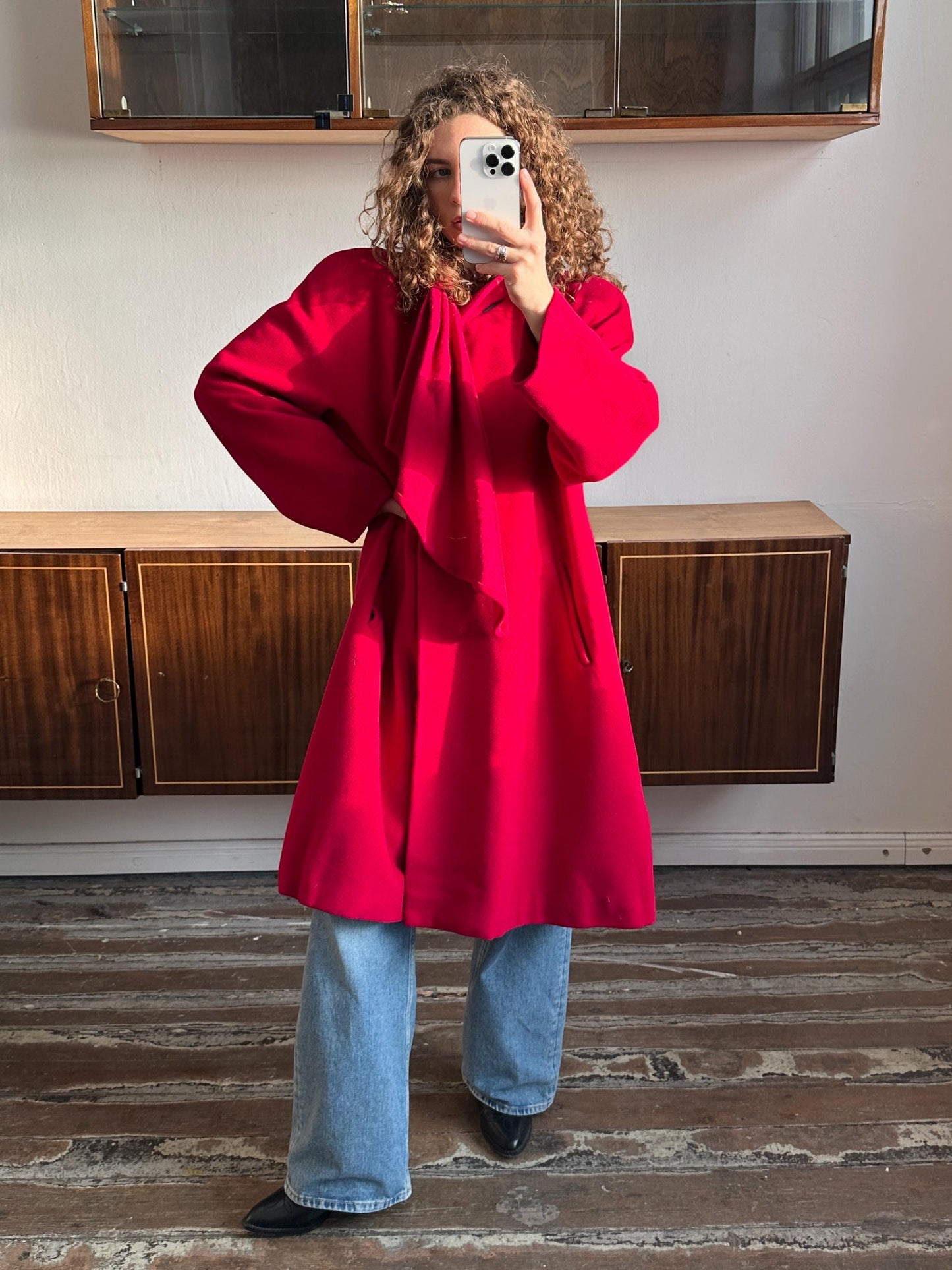 A woman in a red coat is taking a selfie with a smartphone in front of a wooden cabinet.