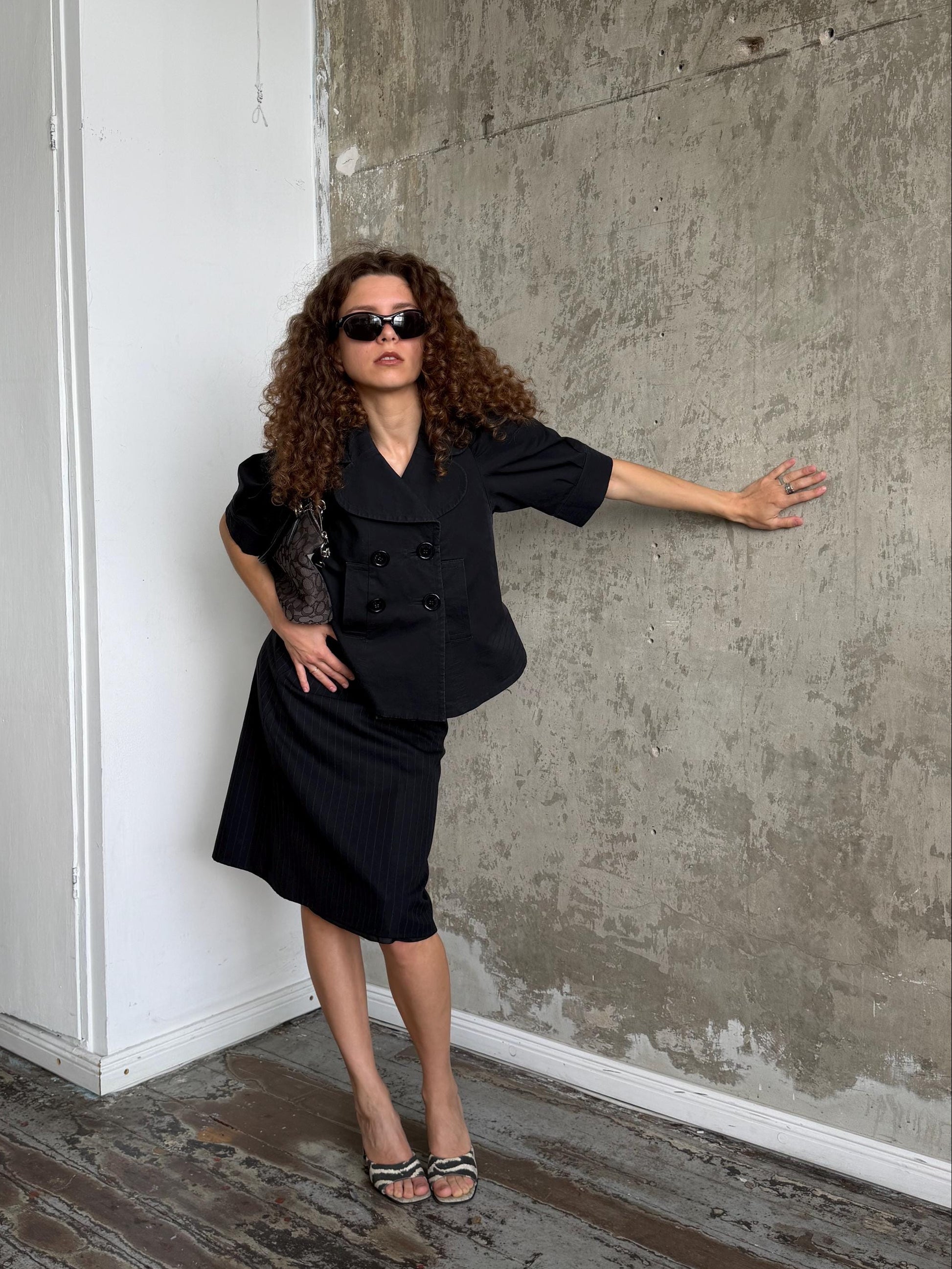 A woman with curly hair wearing a black double-breasted short-sleeve blazer and matching skirt, styled with black sunglasses and heels, posing in a room with a concrete wall and wooden floor.
