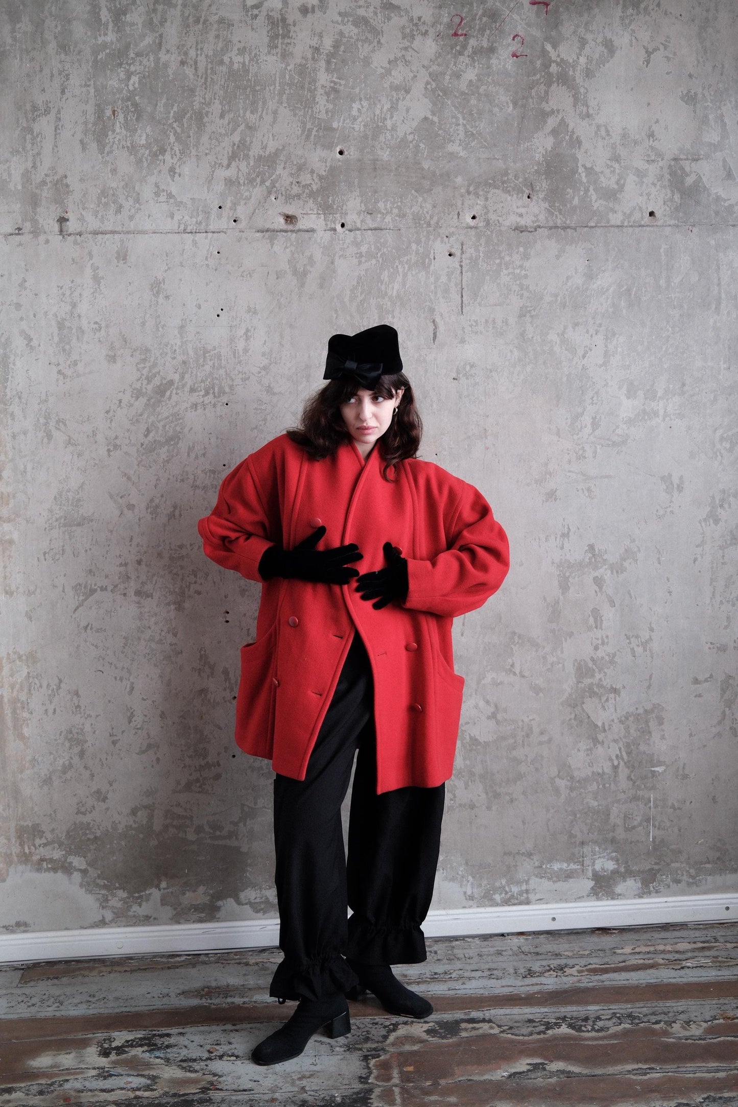 A person wearing a red coat and black pants stands in a room with a concrete wall.