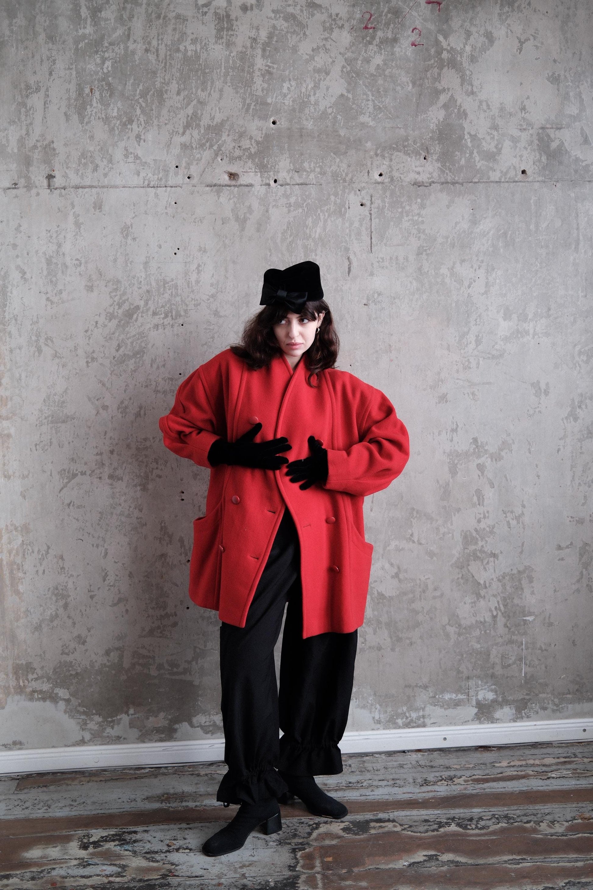 A person wearing a red coat and black pants stands in a room with a concrete wall.