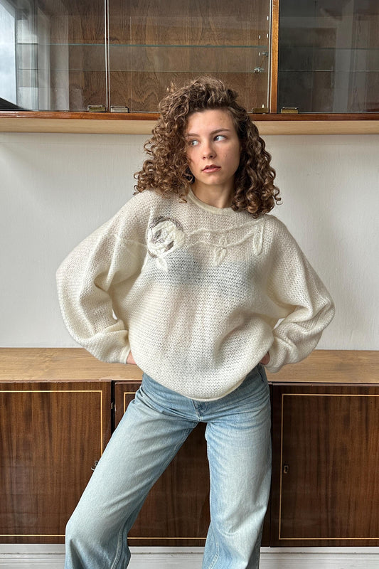 A young woman with curly hair stands in front of a wooden cabinet, wearing a white sweater with a floral design and blue jeans.