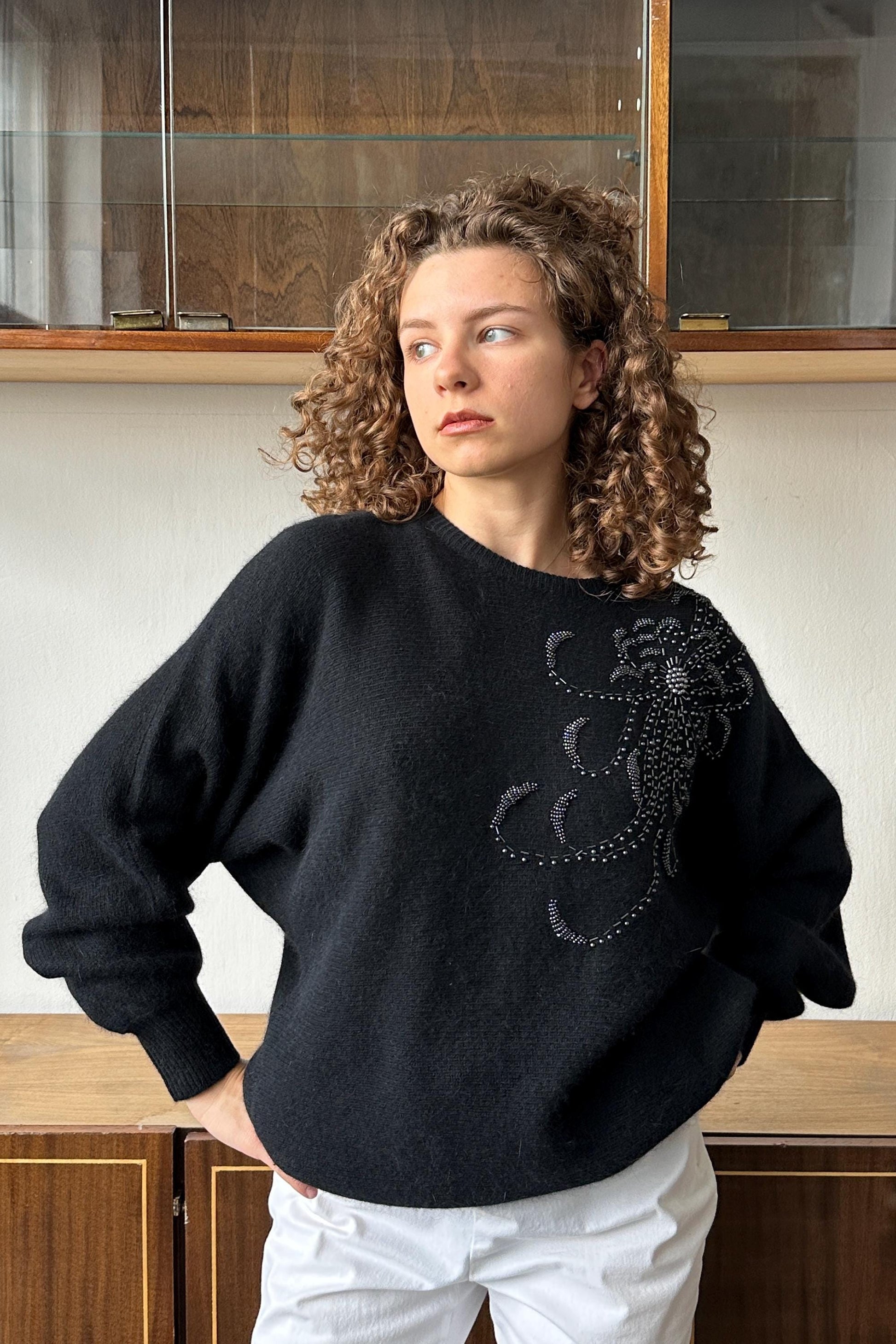 A woman with curly hair wearing a black sweater with a floral embroidered design stands in front of a wooden cabinet.