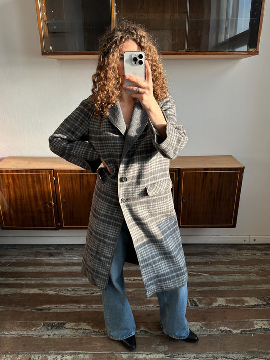 A woman with curly hair is taking a selfie with a smartphone, wearing a gray and black plaid coat and blue jeans.