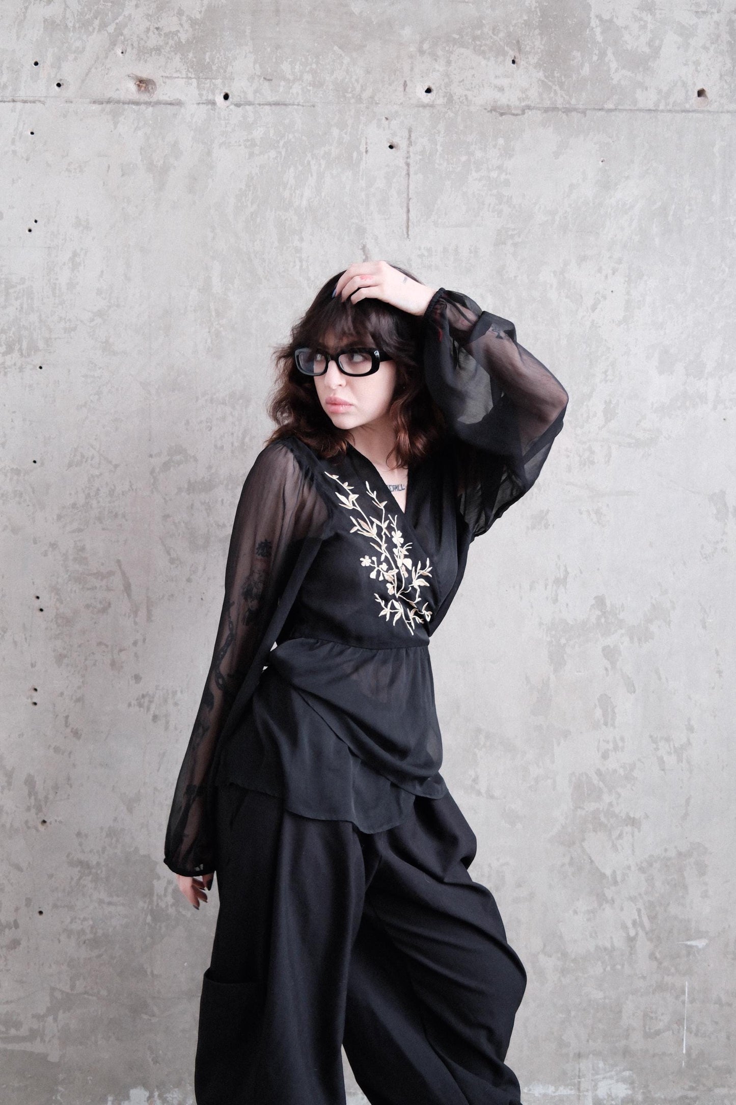 A woman in a black dress with sheer sleeves is lying on a concrete floor, her head resting on her hand.