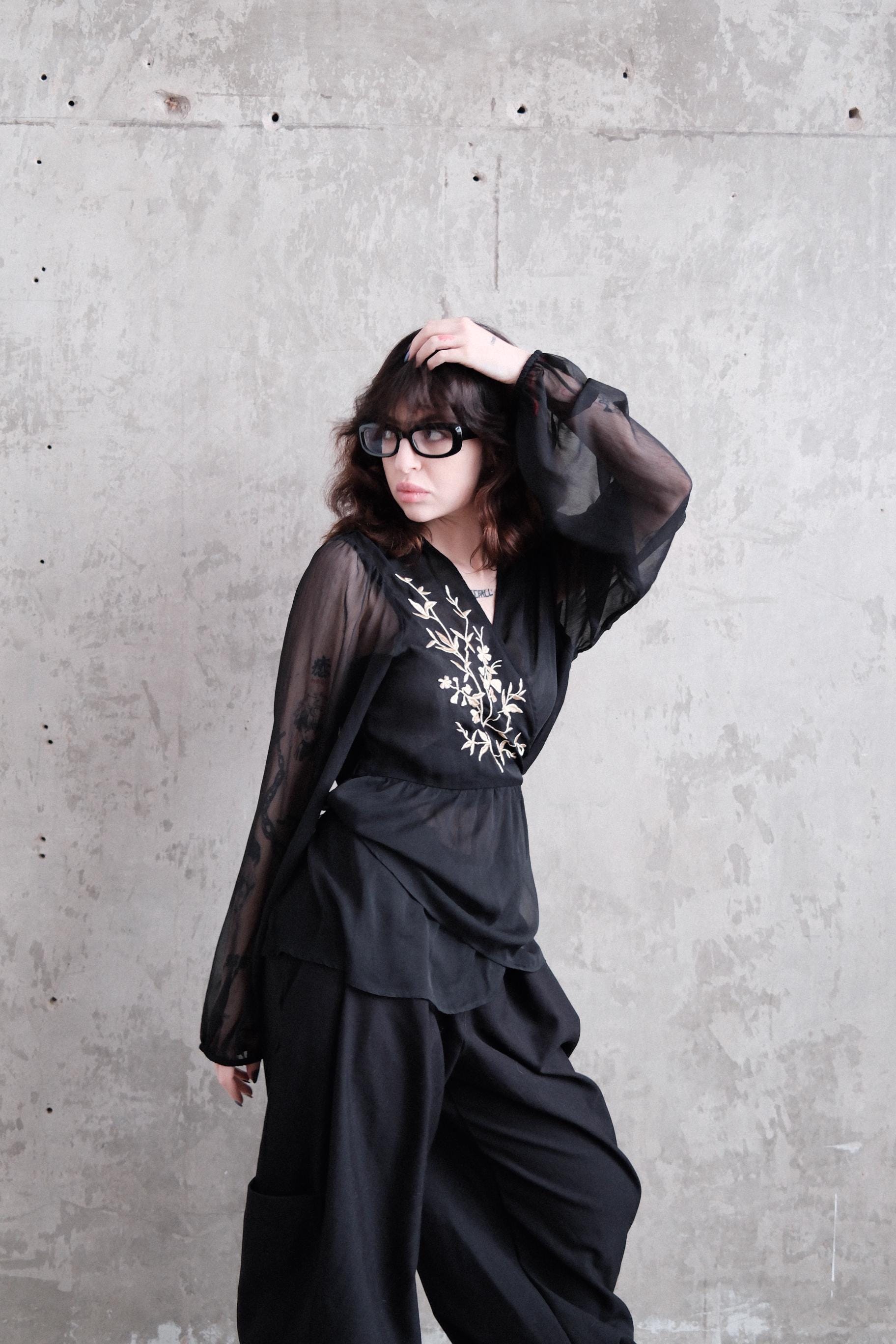 A woman in a black dress with sheer sleeves is lying on a concrete floor, her head resting on her hand.