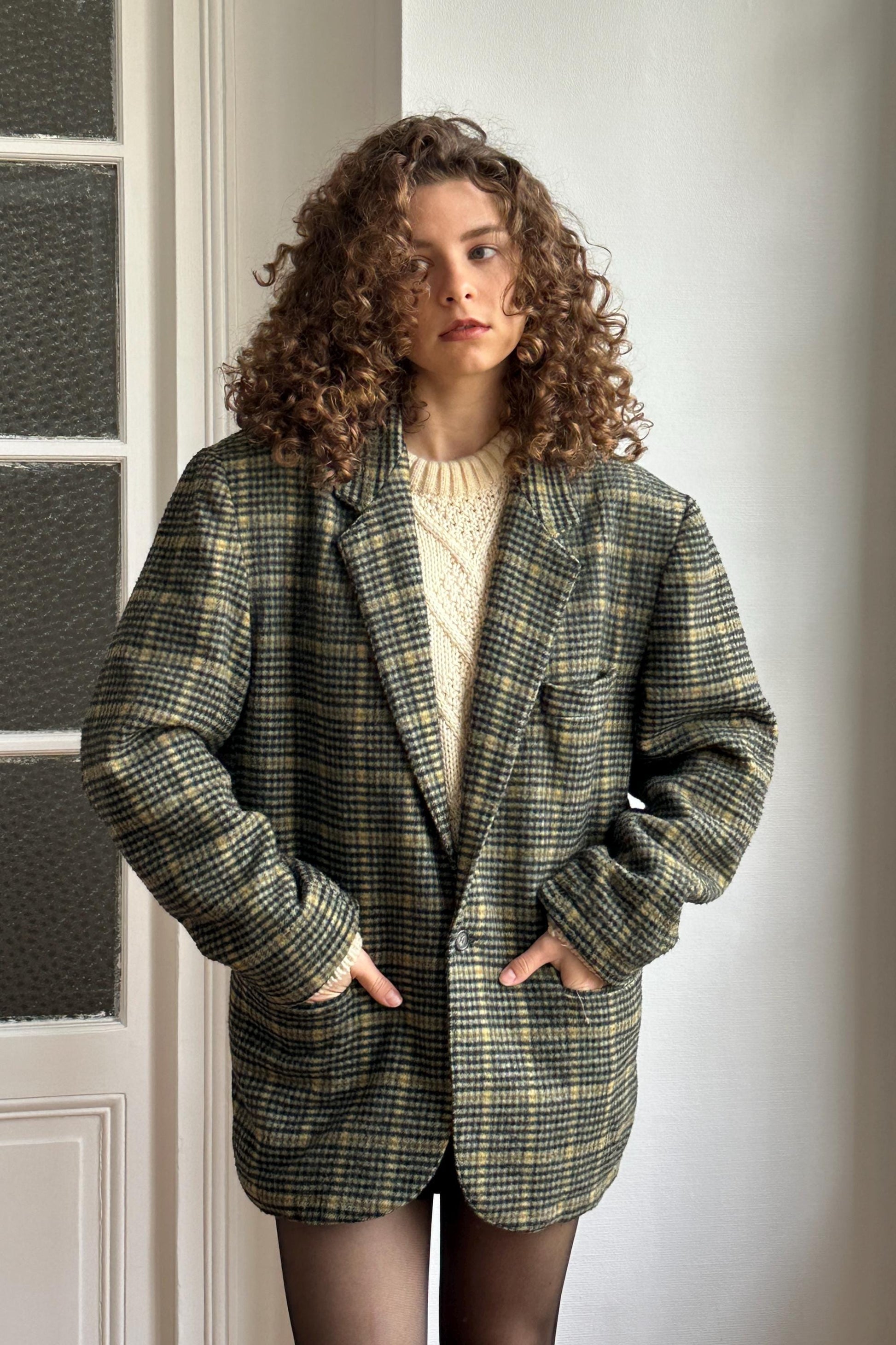 A woman with curly hair wearing a plaid jacket stands in front of a white door.