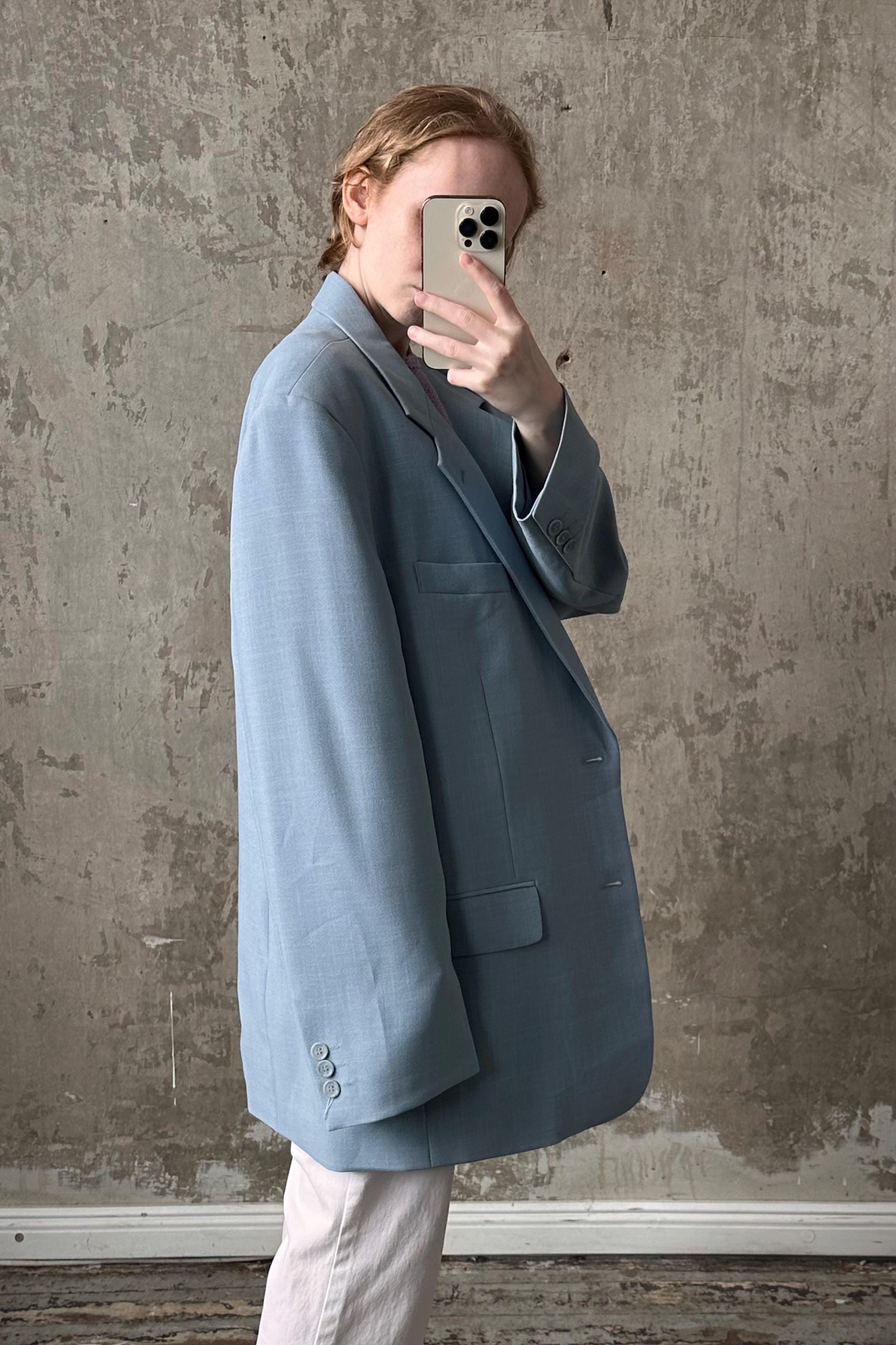 A person is taking a selfie with a smartphone, wearing a light blue blazer against a textured concrete wall background.