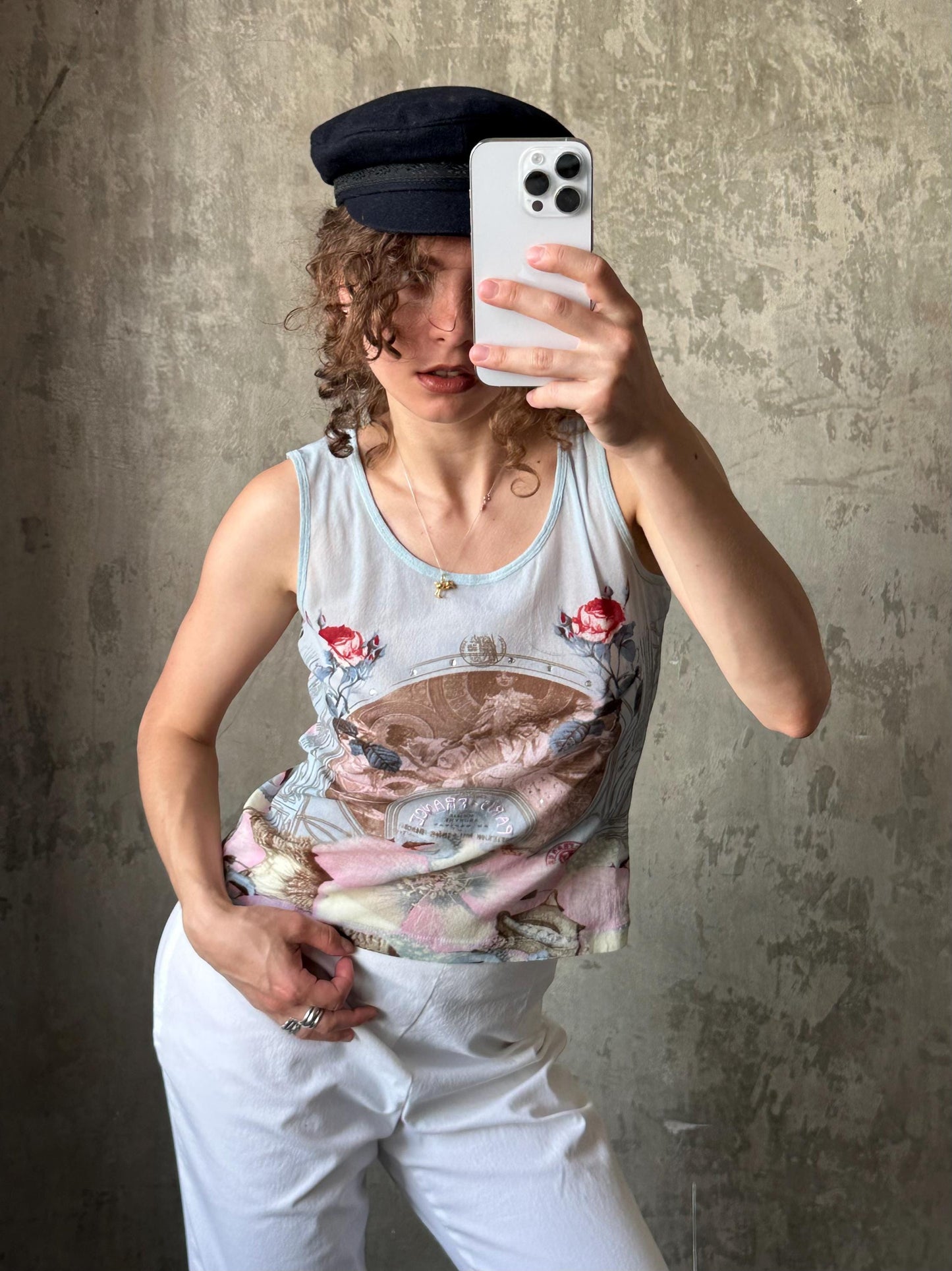 A woman is taking a selfie with a smartphone, wearing a white tank top with a floral design and a black cap.