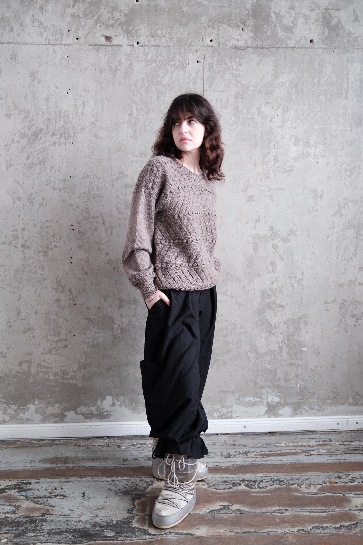 A person wearing a gray sweater and black pants stands in a room with a concrete wall and wooden floor.