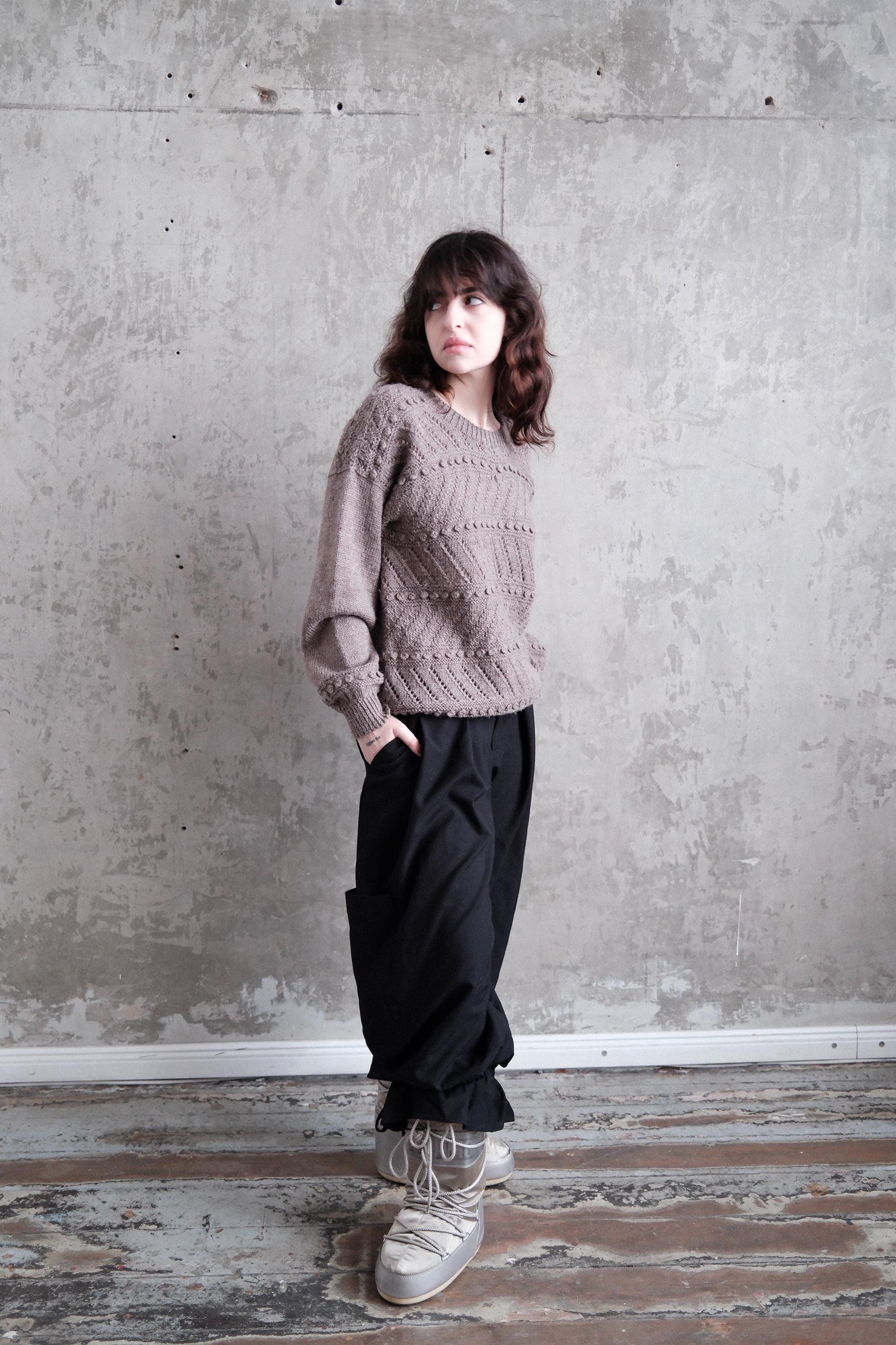 A person wearing a gray sweater and black pants stands in a room with a concrete wall and wooden floor.