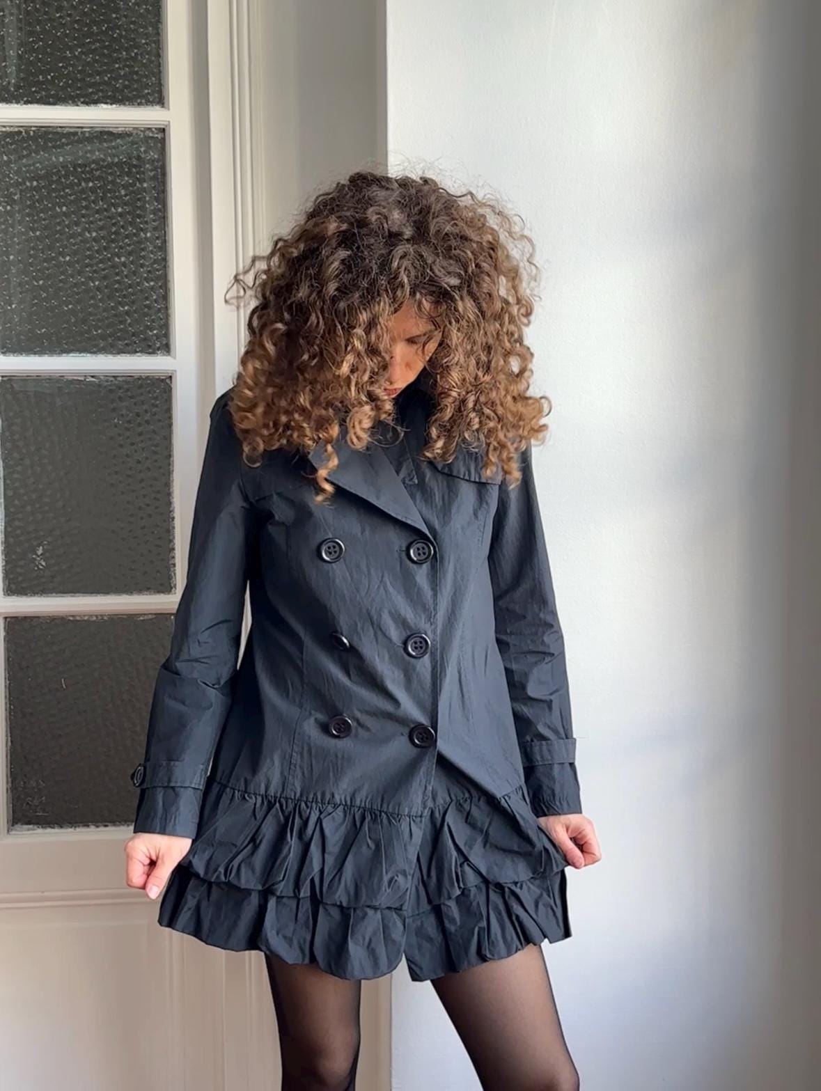 A woman with curly hair wearing a gray coat stands in front of a window.