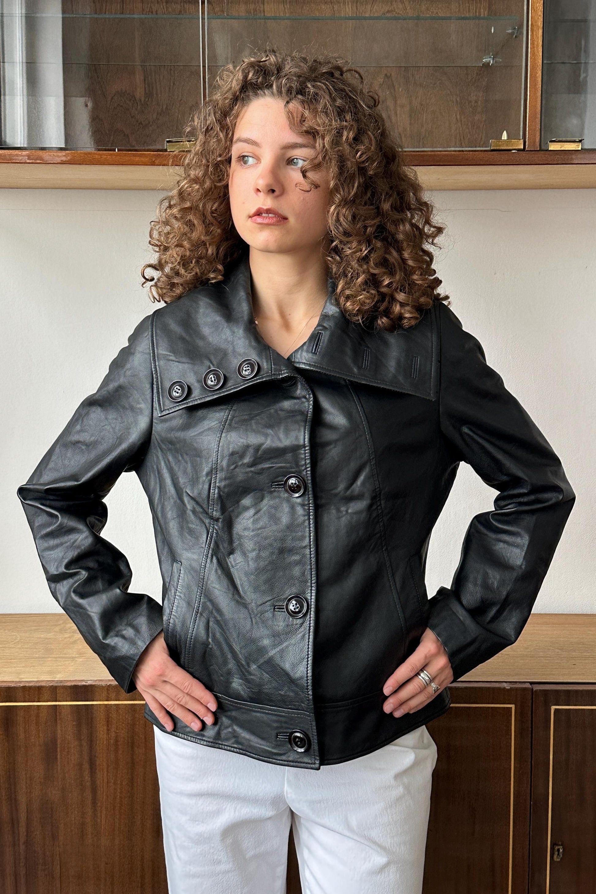 A woman with curly hair wearing a black leather jacket stands in front of a wooden cabinet.