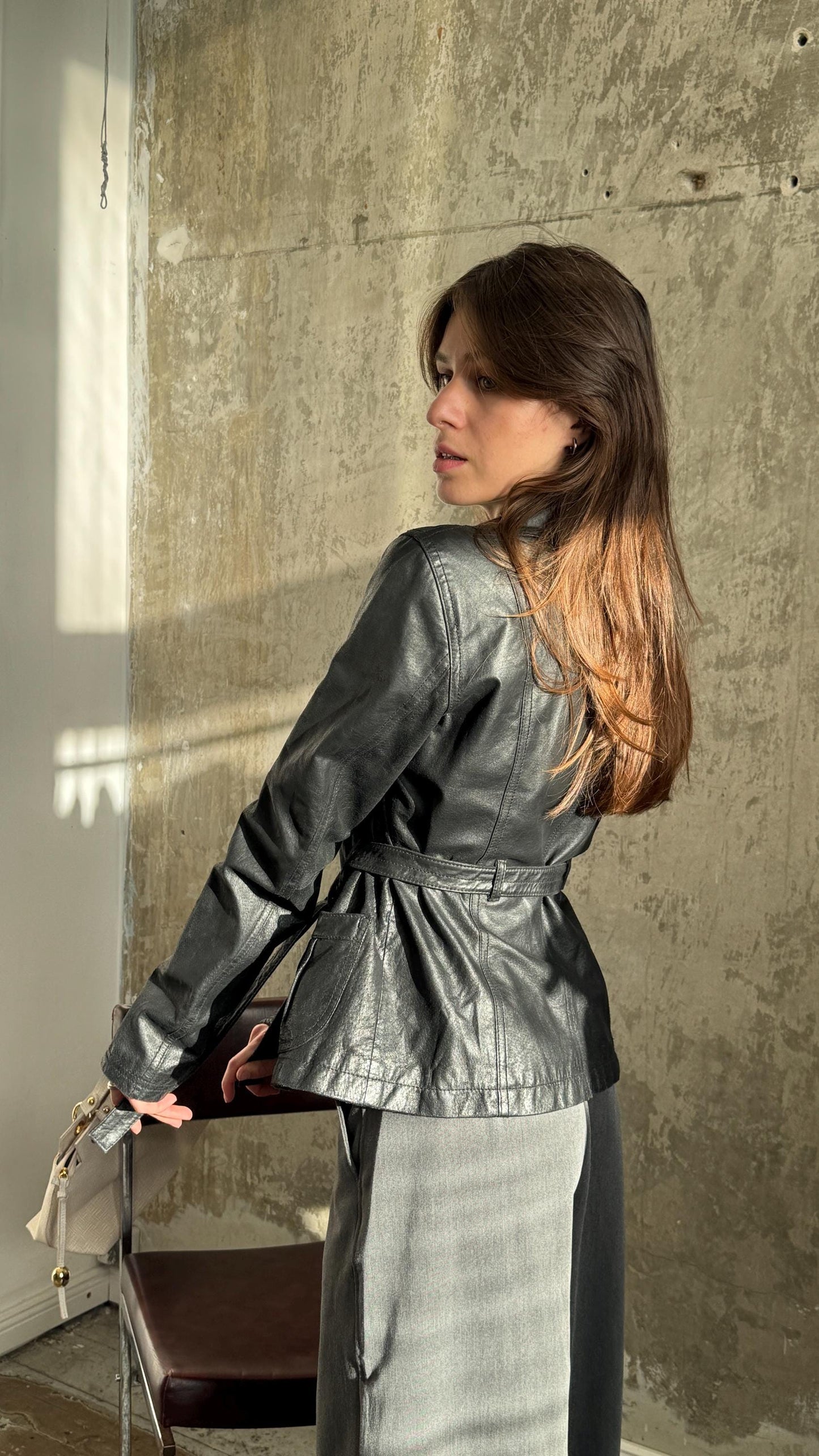 A woman in a black leather jacket stands in front of a concrete wall, her back to the camera.