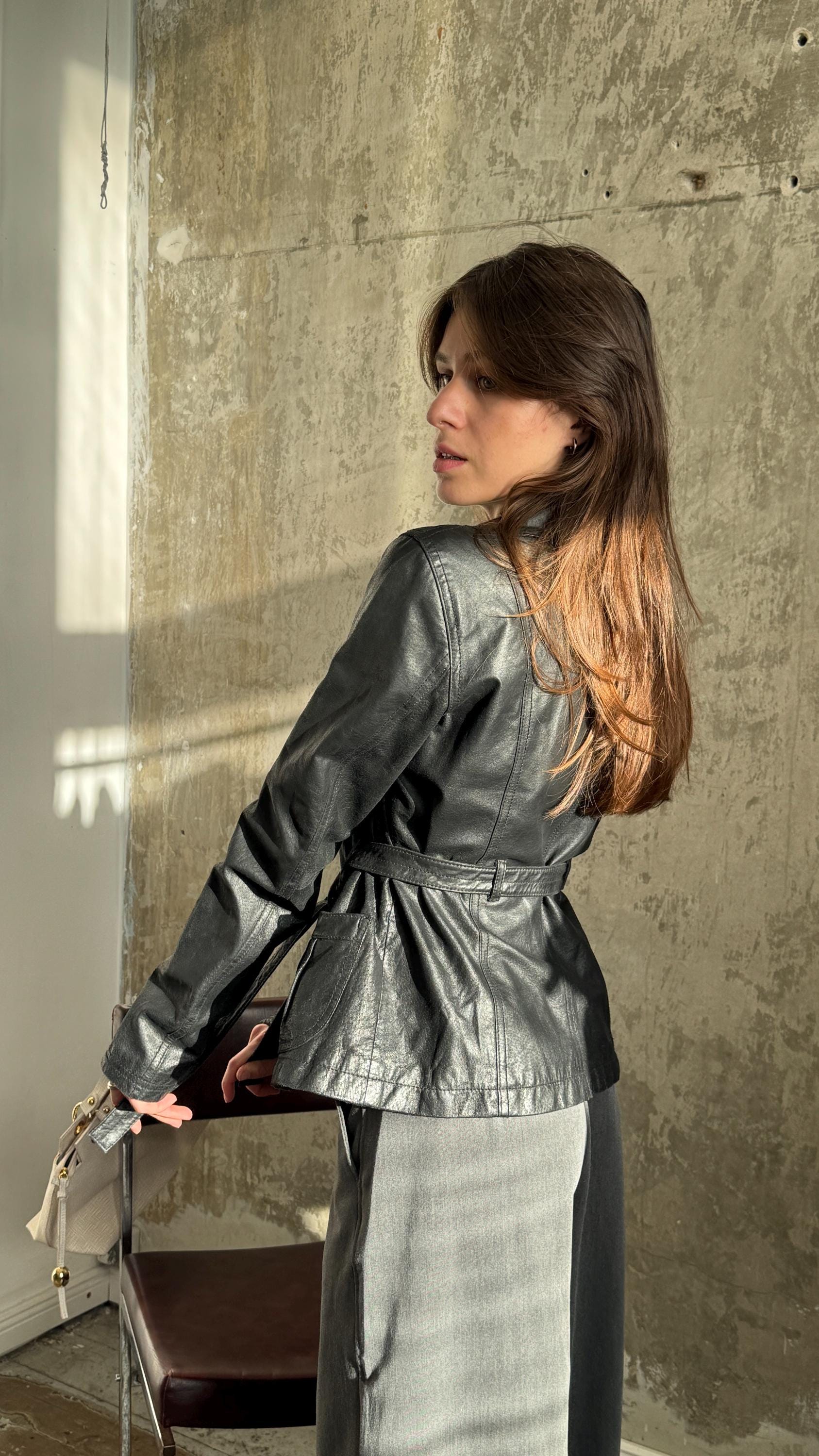 A woman in a black leather jacket stands in front of a concrete wall, her back to the camera.