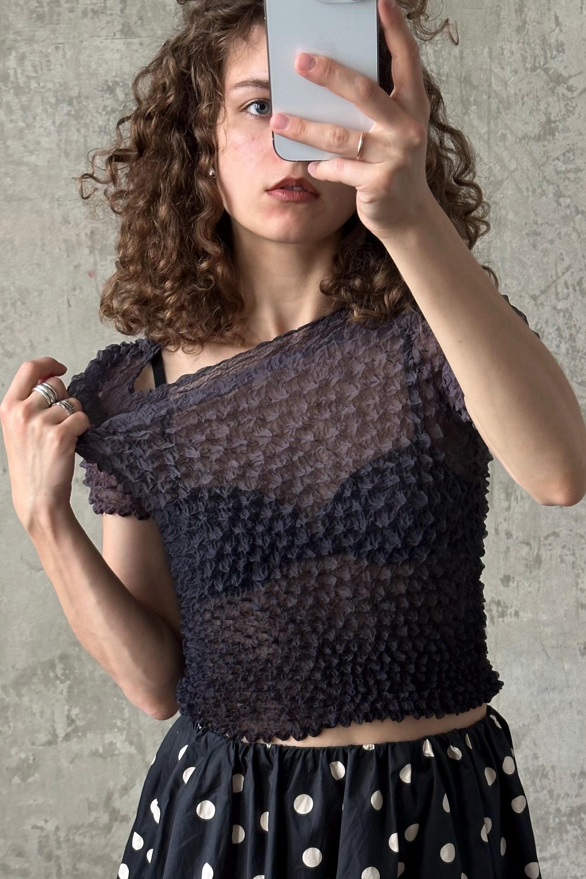 A young woman with curly hair is taking a selfie with a smartphone, wearing a black lace top and a polka dot skirt.