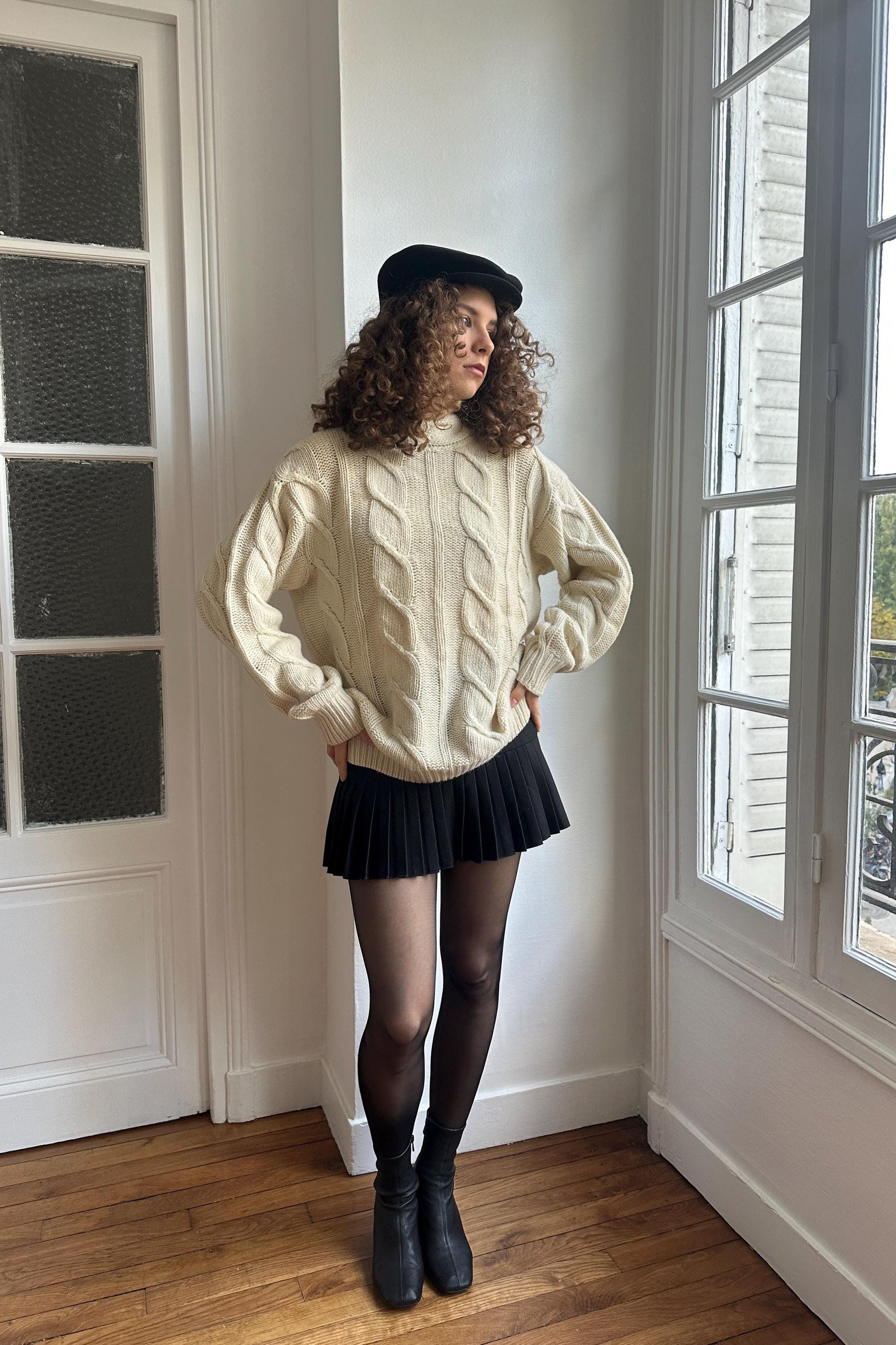 A woman with curly hair stands in a room, wearing a white sweater, black skirt, and black boots. She is looking off to the side, with a window in the background.