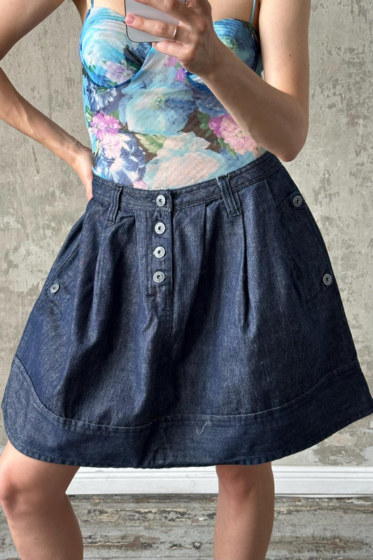 A woman wearing a blue denim skirt and a floral top is holding a phone and appears to be taking a photo or recording a video.