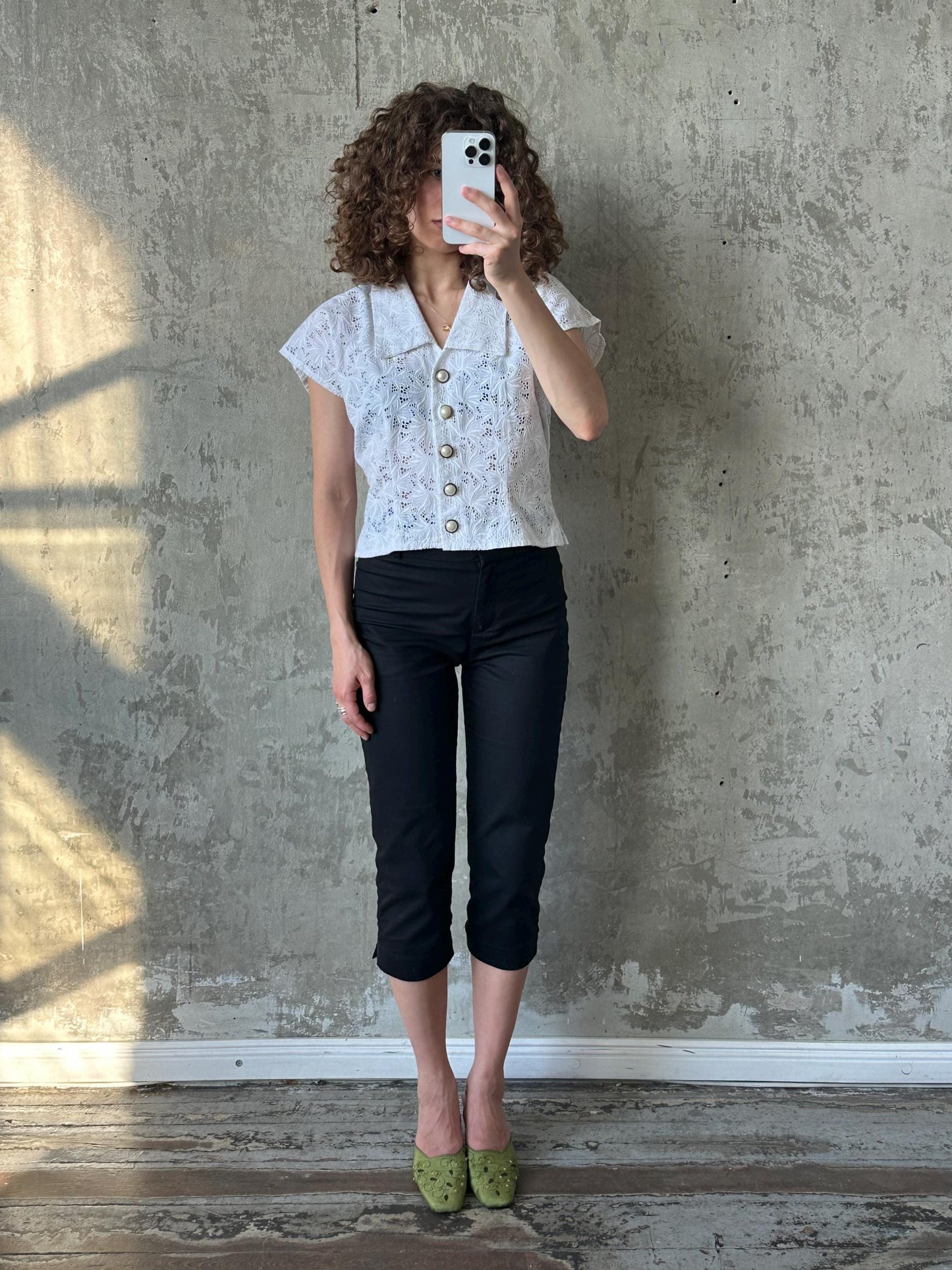 A woman with curly hair is taking a selfie with a white phone, standing against a concrete wall.