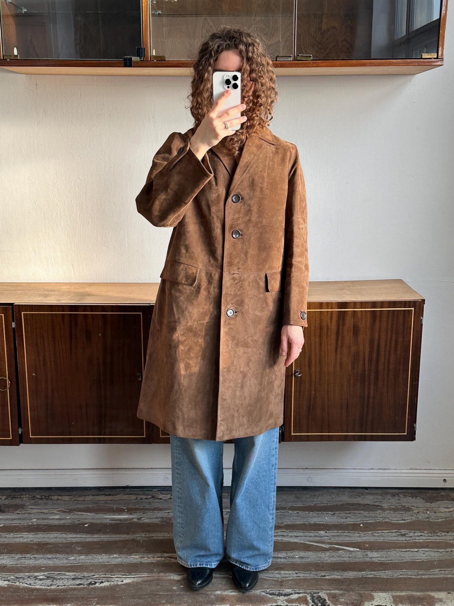 A person is taking a selfie with a brown suede coat and blue jeans, standing in front of a wooden cabinet.
